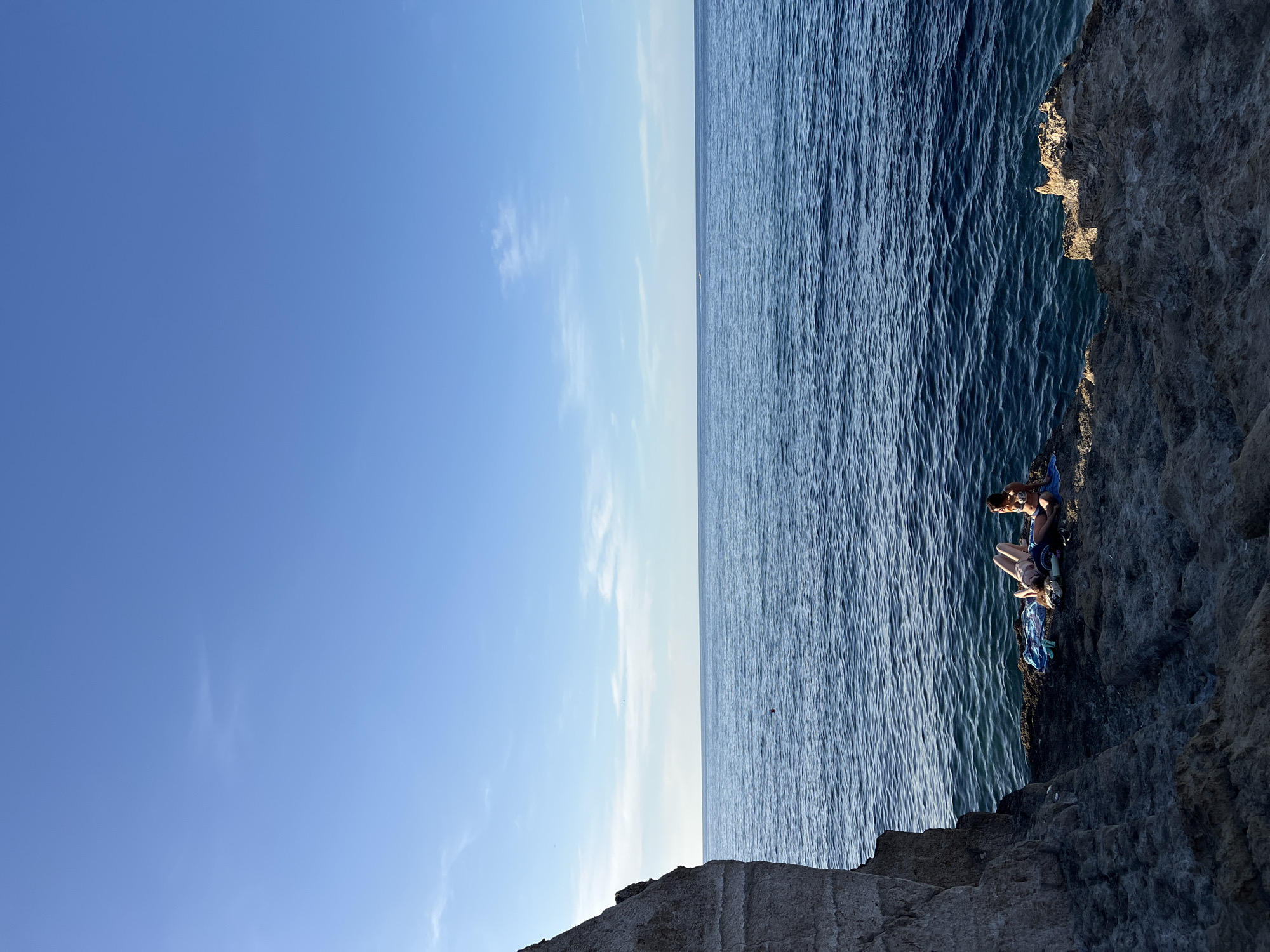 The image depicts a coastal scene in Sperone, Italy, under a clear, bright blue sky with sparse, wispy clouds. An expansive, deep blue sea occupies the mid-ground, meeting a distinct horizon line. Small ripples are visible on the water's surface, and a small, reddish-orange buoy floats in the sea to the left of center. A faint white wake, possibly from a distant vessel, is visible near the horizon.
On the left side of the frame, a large, rugged cliff face, light grey in color and displaying horizontal striations, extends vertically from the bottom edge towards the top. In the foreground, a dark, textured rocky platform extends from the right and foreground towards the cliff and the sea.
Two adult females are situated on a flatter section of this rocky platform, adjacent to the water's edge. Both are wearing swimwear. The female on the left is lying supine on a blue and white striped towel, facing upwards, with her legs slightly bent. The female on the right is seated upright on a blue towel, facing towards the water or slightly towards the first individual. Dark footwear is visible near her feet on the towel. Both individuals appear to be relaxing and sunbathing. The scene is brightly illuminated by natural light, casting subtle shadows on the rocky formations.