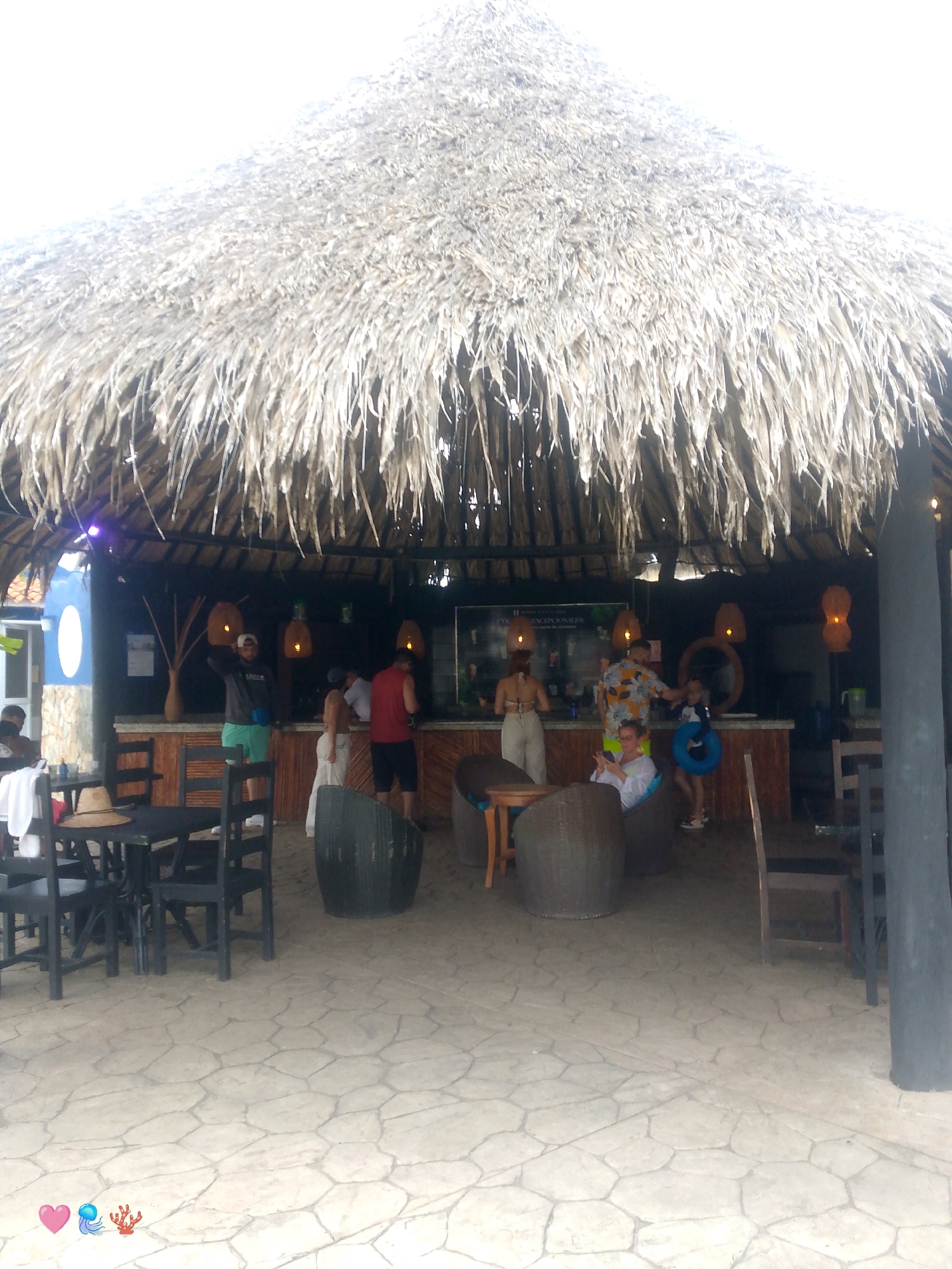 The image depicts a scene at a beachside bar in Playa El Agua, Venezuela. A prominent thatched roof structure covers an open-air bar area. Several people are visible in and around the bar, some standing and others seated. The floor is paved with large, irregular stones. Tables and chairs, some made of dark wood and others of woven material, are placed around the seating area. In the background, the bar itself is visible with bottles and other items on display. Decorative pendant lights hang from the ceiling. The overall atmosphere appears casual and relaxed, typical of a beach resort setting.