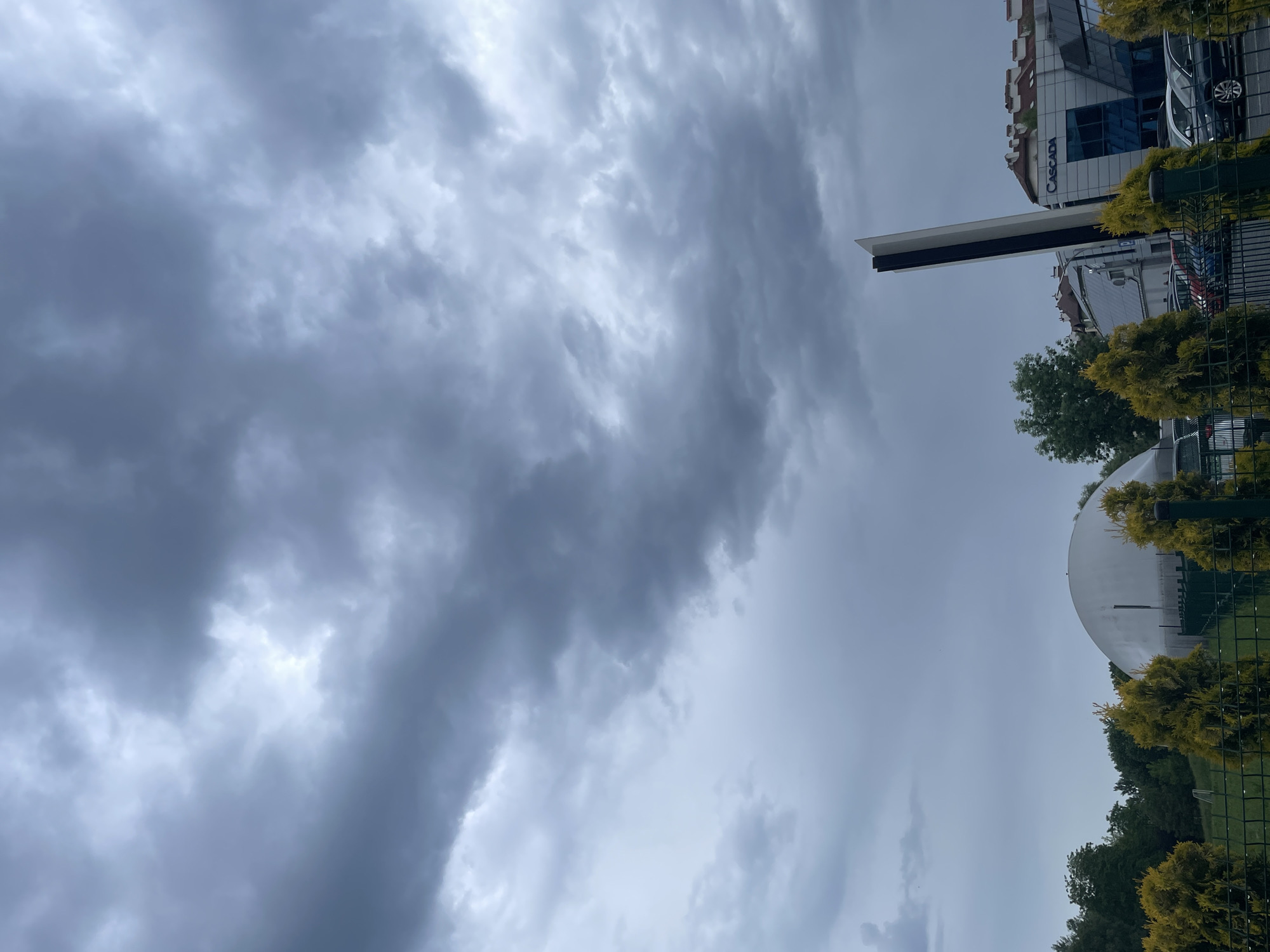 An overcast sky filled with dark grey and lighter grey clouds dominates the upper and central portions of the image, suggesting a heavily clouded or pre-stormy day. Below the sky, an urban or suburban landscape is visible.
On the right side of the frame, a multi-story building with reflective windows is partially seen, featuring a prominent blue sign that reads "CASCADA." In front of this building, a tall, slender, rectangular structure, predominantly white with a black section, stands upright. Further to the right, parts of another building and a dark-colored car are visible, suggesting a parking area or street.
Towards the lower right, a large, smooth, white, curved structure, possibly a dome or a large satellite dish, is partially obscured by trees. The foreground features a line of bright yellow-green, manicured bushes or conifers, running alongside a green metal fence. Beyond this fence, a dense cluster of dark green trees is visible, indicating more natural vegetation.
The scene is static with no discernible human activity. The diffused light suggests it is daytime under heavy cloud cover.