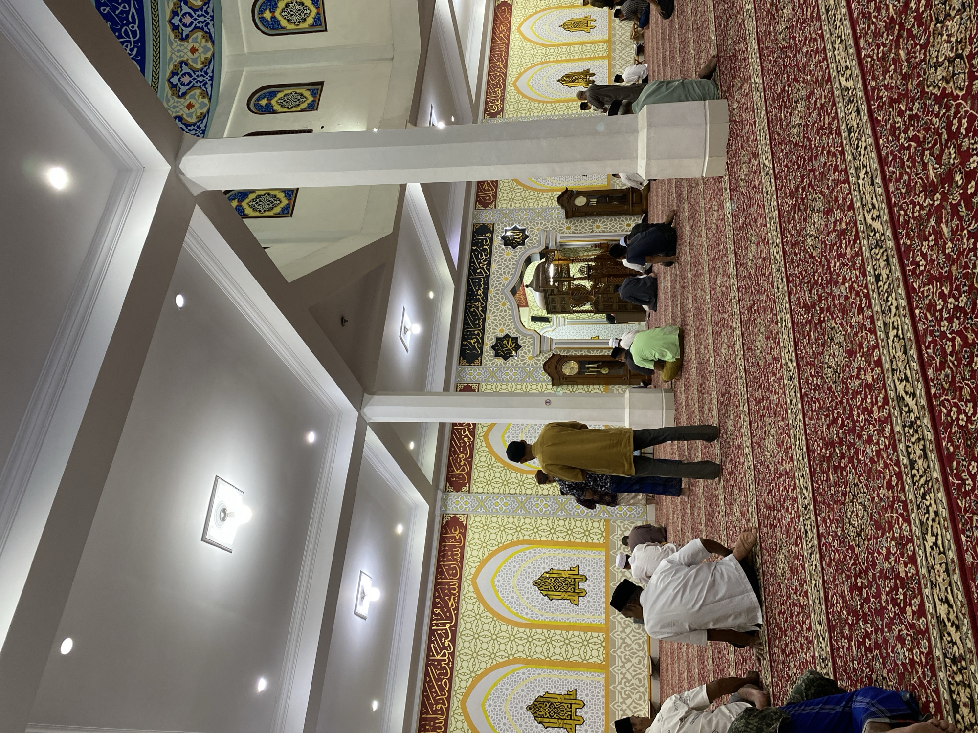 The image depicts the interior of a mosque in Lhokseumawe, Indonesia, filled with worshippers seated on a red carpet with intricate gold and black patterns. The mosque features high ceilings with modern lighting fixtures, large white pillars, and ornate yellow walls decorated with Islamic calligraphy and geometric designs. In the background, a detailed wooden pulpit is visible, flanked by antique clocks. Several men are seen in prayer, with some standing and others kneeling. The overall ambiance is one of solemnity and community gathering.