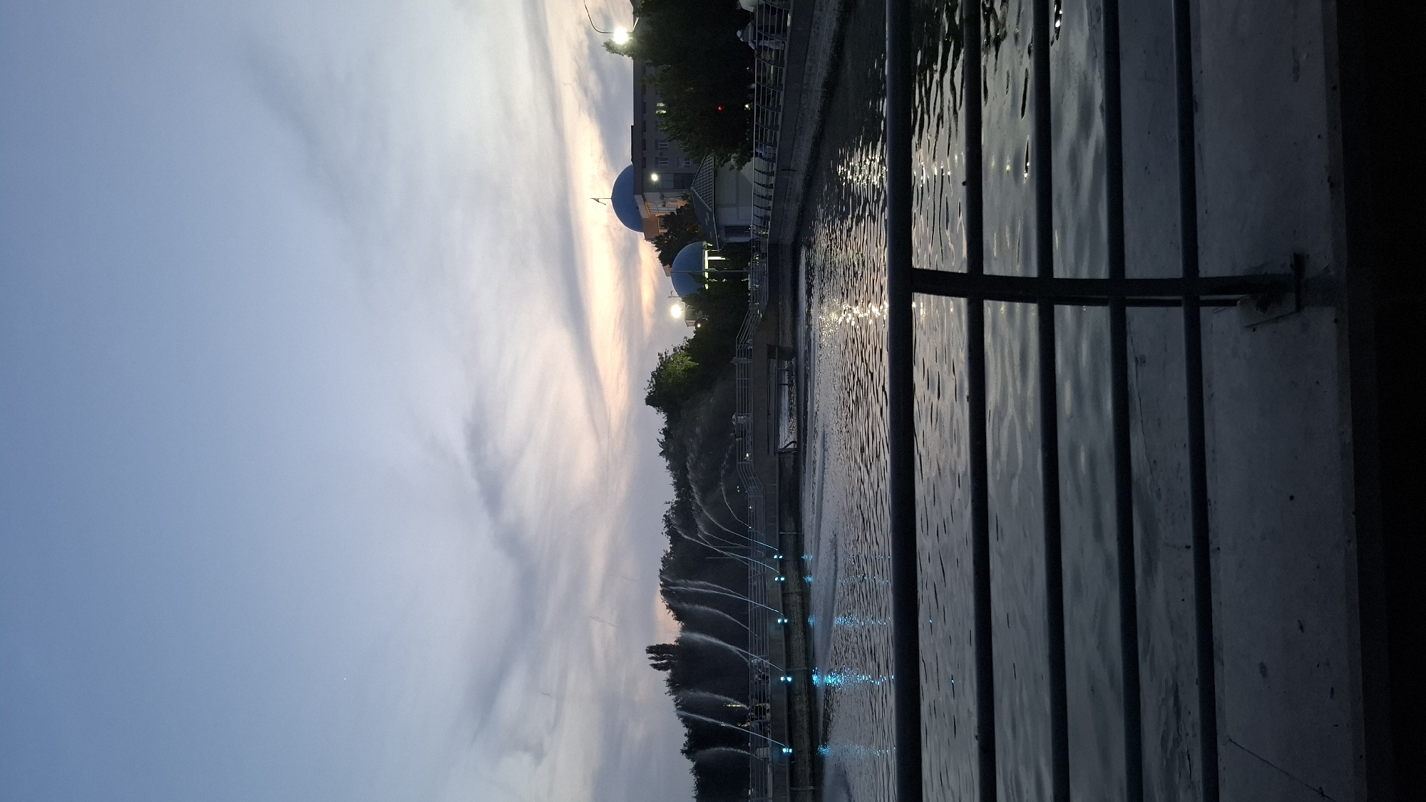The image depicts a public park at dusk, likely located in Shymkent, Kazakhstan. In the foreground, a dark metal railing separates the viewer from a body of water. The water's surface is rippled, reflecting the dim light of the sky. Across the water, a row of illuminated fountains sprays water upwards, creating arcs of light. Beyond the fountains, a tree-lined area is visible, with buildings and lights suggesting an urban setting. Two domed buildings, one with a flag, are particularly prominent. The sky is a gradient of pale blue and grey, with streaks of pink and orange indicating the setting sun. The overall atmosphere is serene and peaceful.
