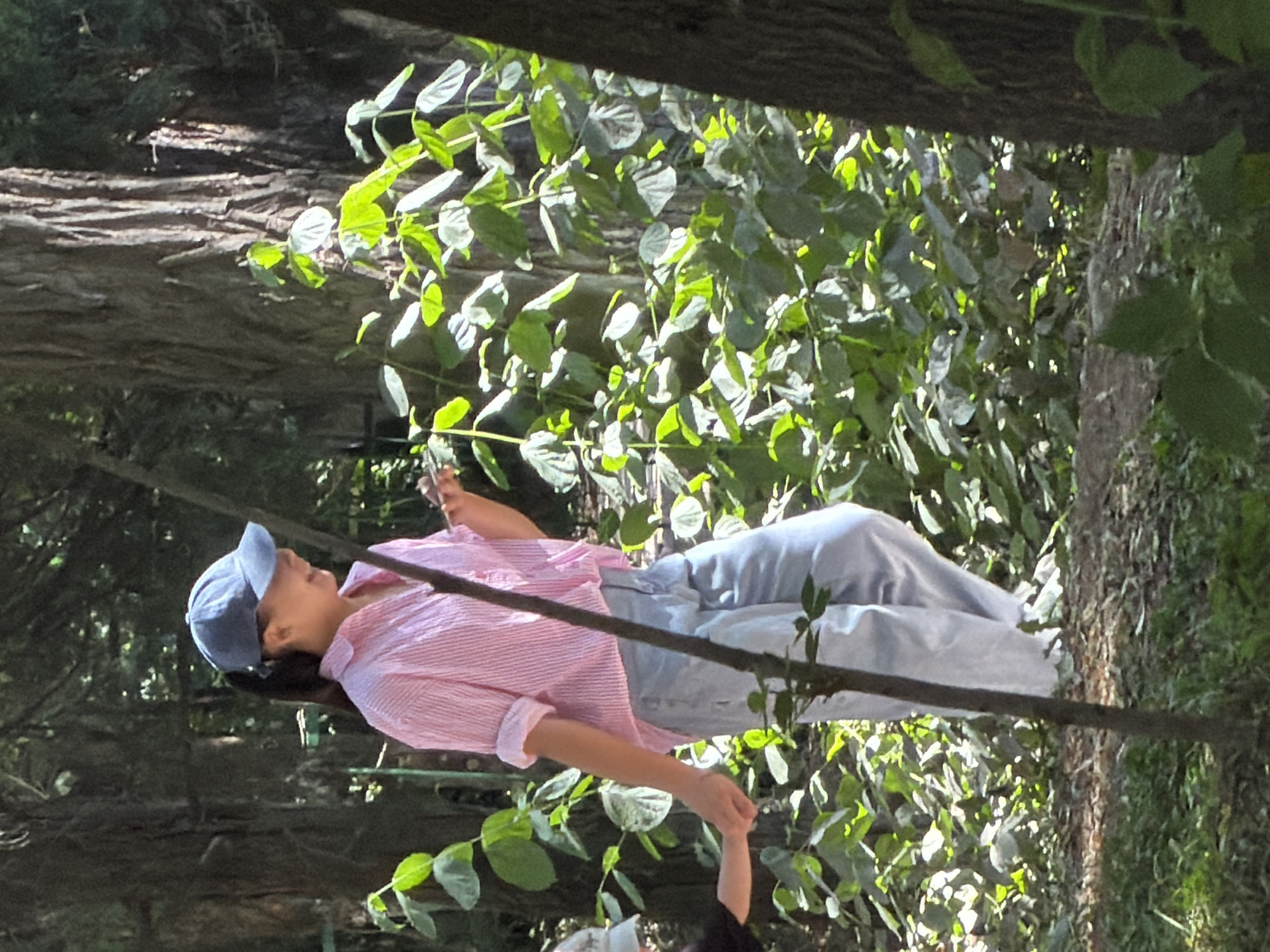 A young woman wearing a pink and white striped shirt, light blue pants, and a blue baseball cap is walking in a wooded area and holding hands with a child. The woman is smiling and looking down. The child's face is not visible. The environment is a forest with tall trees and lush green foliage. The ground is covered with dirt and leaves. The image appears to be taken during the day, with sunlight filtering through the trees.