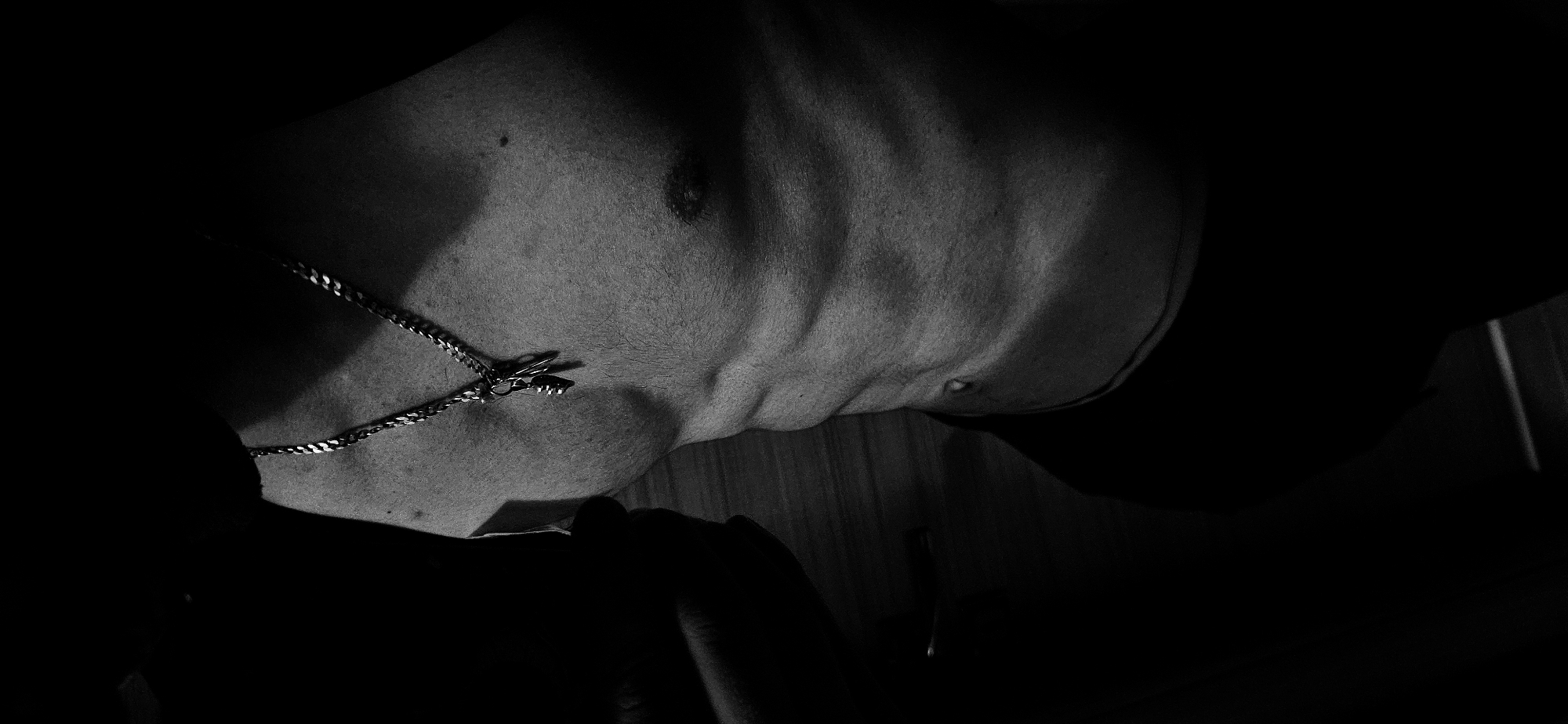 A close-up, black-and-white image shows the torso of a person with defined musculature, including the clavicle and abdominal area. The person is wearing a silver necklace with a clasp visible. Skin details such as pores and body hair are noticeable. The lighting is dramatic, casting strong shadows and highlighting muscle contours. The lower portion of the image is obscured by deep shadow, making it difficult to identify specific objects. The scene does not show any details that could indicate a location in Plovdiv, Bulgaria.