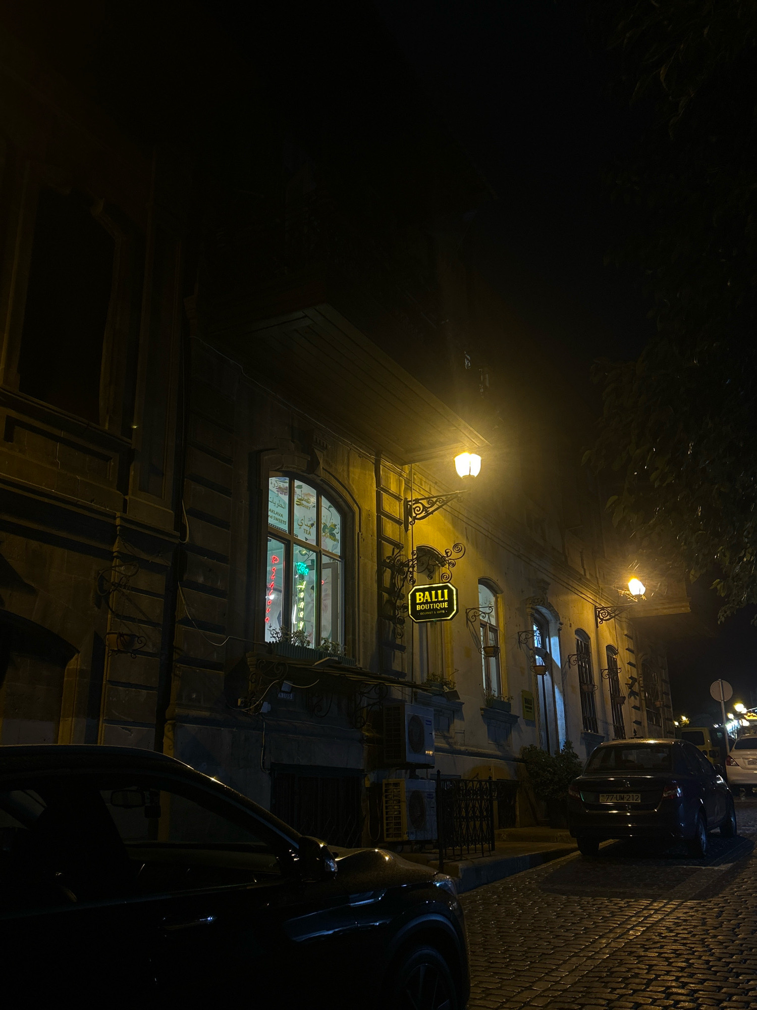 The image captures a nighttime scene on a cobblestone street in Sabail Raion, Azerbaijan. A dark-colored sedan is parked on the right side of the street, facing away from the viewer. Another dark vehicle is partially visible in the foreground on the left. The buildings lining the street are illuminated by warm-toned streetlights and lights emanating from windows. One building features a sign that reads "BALLI BOUTIQUE GOURMET & COFFEE." The window of a nearby establishment displays neon signs, one of which appears to be in an Arabic script. The street is paved with cobblestones, and trees with dark foliage overhang the scene. The overall atmosphere is quiet and dimly lit, typical of a city street at night.