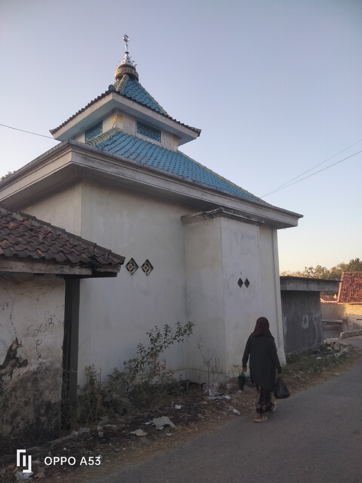 A full-shot, eye-level outdoor photograph captures a mosque with a blue tiled roof and a crescent moon finial on its dome. The mosque is situated next to a smaller, dilapidated building with a tiled roof. A woman wearing a hijab and a dark jacket walks away from the mosque on a paved road, carrying two plastic bags. She is dressed in patterned pants and light-colored shoes. The environment appears to be a rural or semi-rural area, with sparse vegetation and a clear sky. The image is taken from the back of the woman, looking towards the mosque and the road. The overall lighting suggests it is either early morning or late afternoon. The OPPO A53 logo is visible in the bottom left corner.