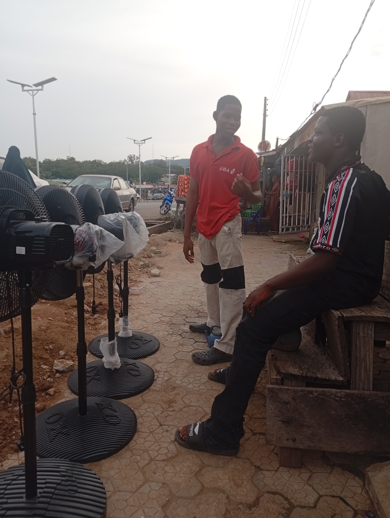 In Zango-ndaji, Nigeria, two young men stand beside a row of black standing fans displayed for sale on a tiled pavement. The man on the left, wearing a red polo shirt with a "UBA" logo and beige pants with black patches, gestures with his left hand while speaking to the man on the right. The second man, dressed in a black t-shirt with a geometric pattern on the sleeves and black pants, is seated on a rough wooden bench. He appears to be listening to the conversation. Behind them, a street scene unfolds with a silver car, a blue motorcycle, and other individuals in the background, suggesting a busy marketplace or roadside commercial area. The environment features overhead utility lines and streetlights, indicating an urban setting. The fans, some covered in plastic, are arranged in a line, their bases resting on the patterned tiles. The ground around the pavement is a mix of dirt and gravel.