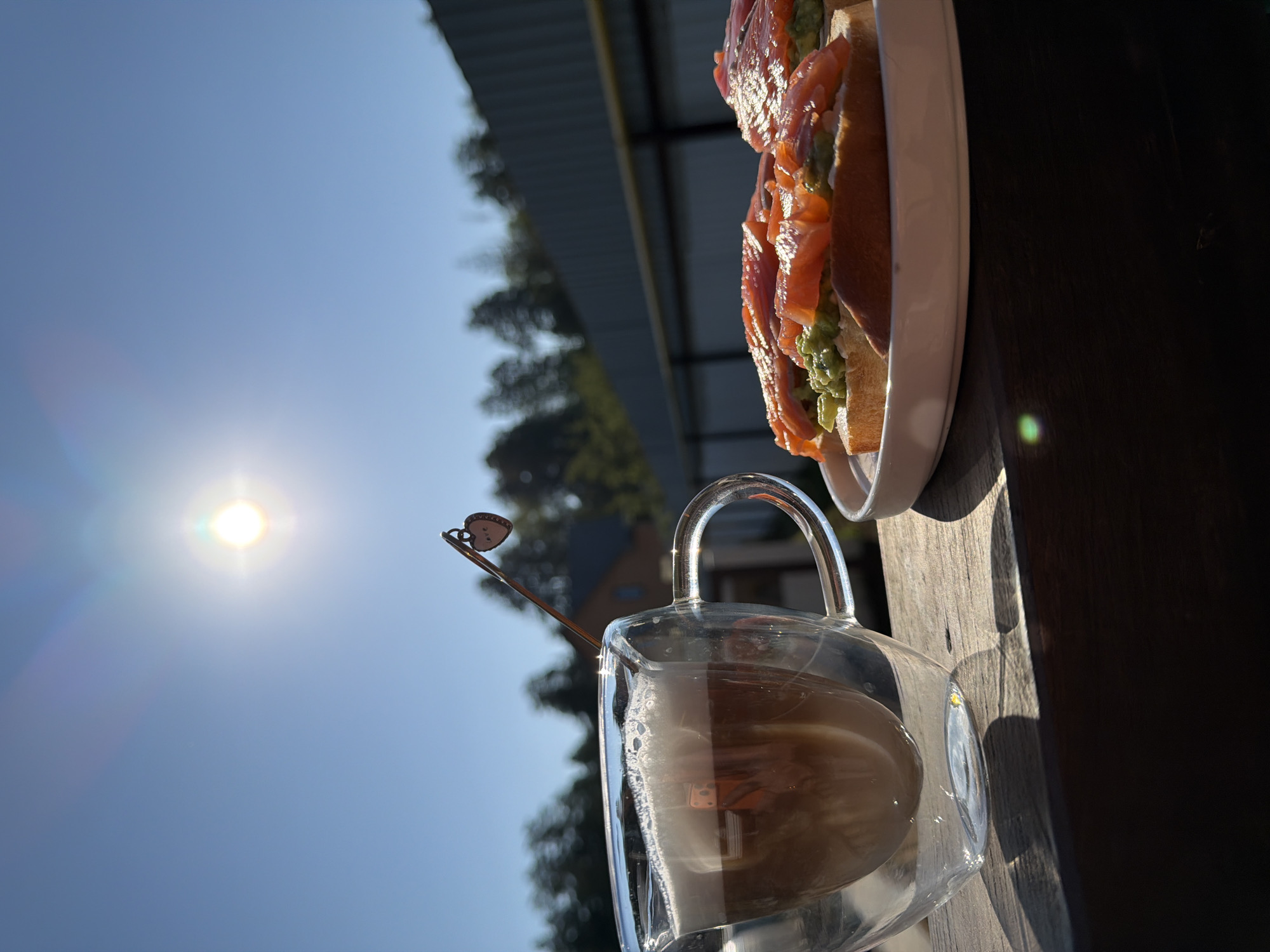 The image shows a still life composition. A clear glass mug with a beverage inside is placed on a wooden surface, likely a table. A metal spoon with a decorative charm hangs on the rim of the mug. Next to the mug, a white plate holds a piece of toast topped with avocado and salmon. The background features a clear blue sky with a bright sun. There are trees visible behind the plate, and a portion of a building structure is visible. The image is rotated counter-clockwise. The image appears to be taken outdoors, potentially at a restaurant or cafe in Chaikovsky, Russia.