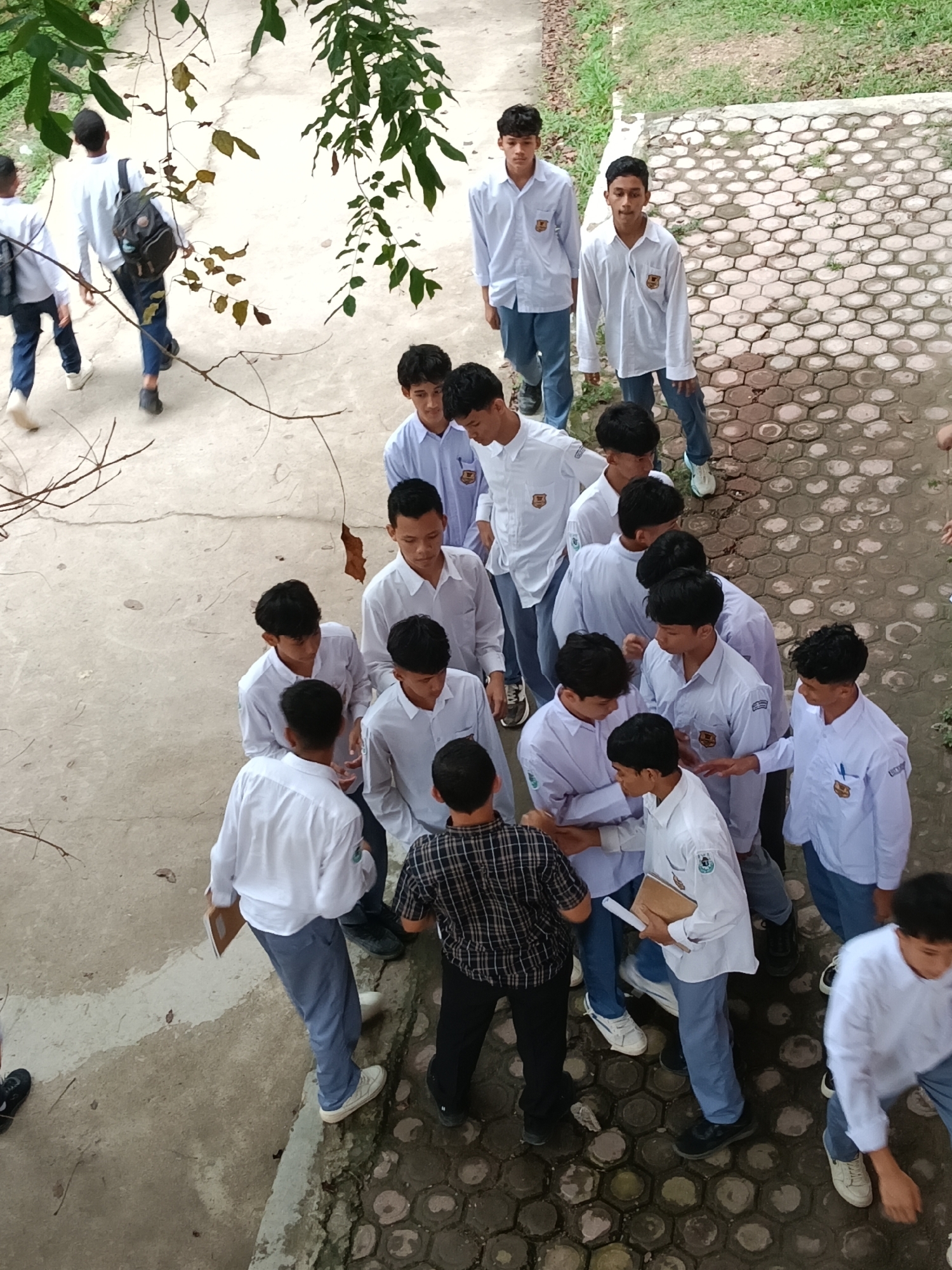 From an elevated vantage point, the image captures a gathering of male students in a school setting, likely located in Lhokseumawe, Indonesia. The students are predominately wearing white shirts and blue/gray trousers, presumably the school uniform. A group of students are clustered closely together, while some are standing at the periphery or walking away. One student wears a black and white checkered shirt, standing among the uniformed students. A student near the group holds books and papers. The background features a combination of concrete and paved areas, along with a few patches of greenery. Overhead, there are tree branches with leaves visible. The image appears to be taken outdoors during the day.