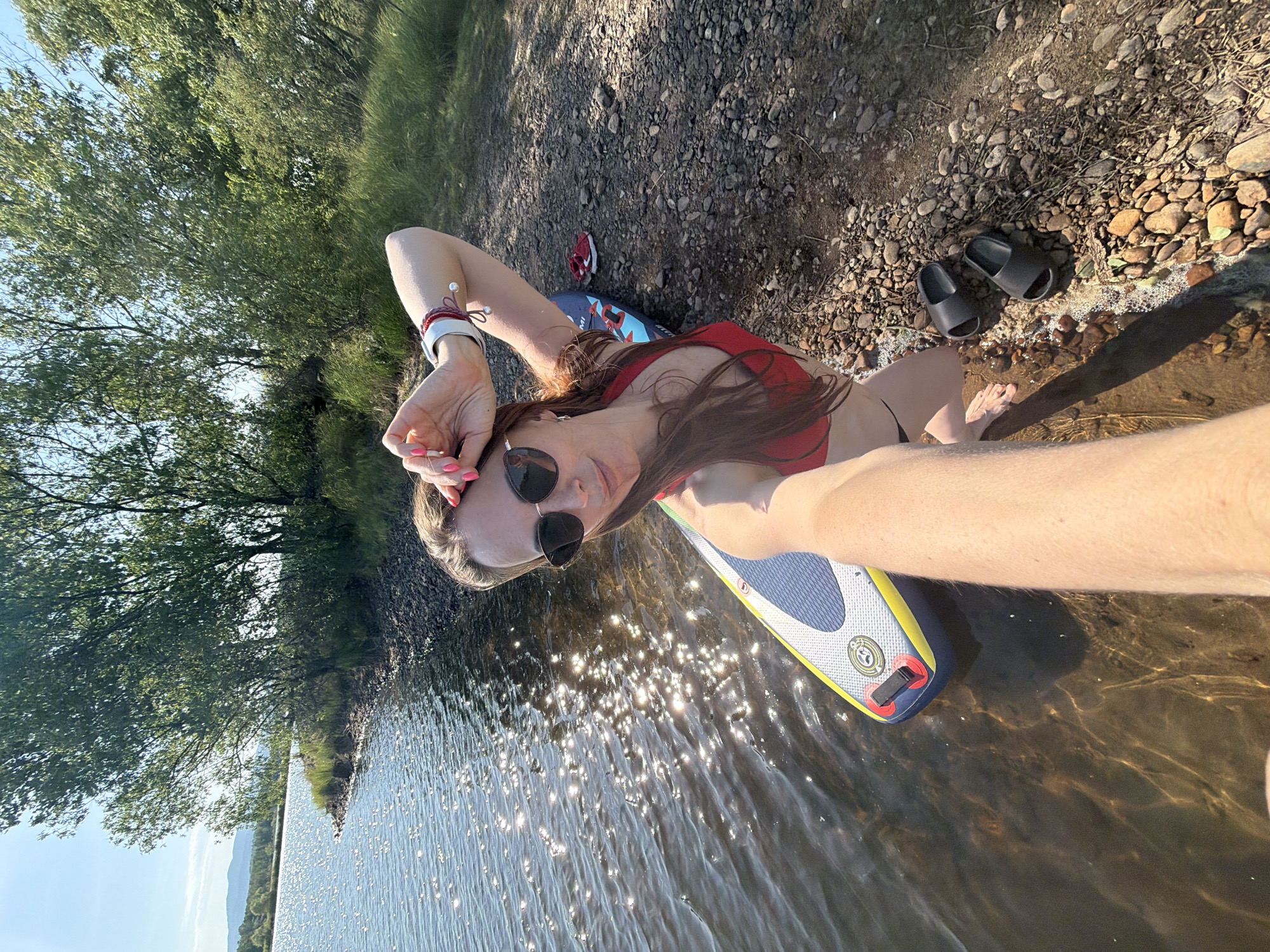 The image depicts a woman in sunglasses and a red bikini lying on a paddleboard at the edge of a body of water. Her arm is raised, possibly in a selfie pose. The paddleboard is partly submerged in the water, which reflects sunlight. A tree line is visible on the shoreline, with rocks and pebbles in the foreground next to the board. A pair of black slides and a red and white sneaker are also on the shore. The location appears to be a lake or riverbank. The image does not provide enough information to determine if it was taken in Birobidzhan, Russia.