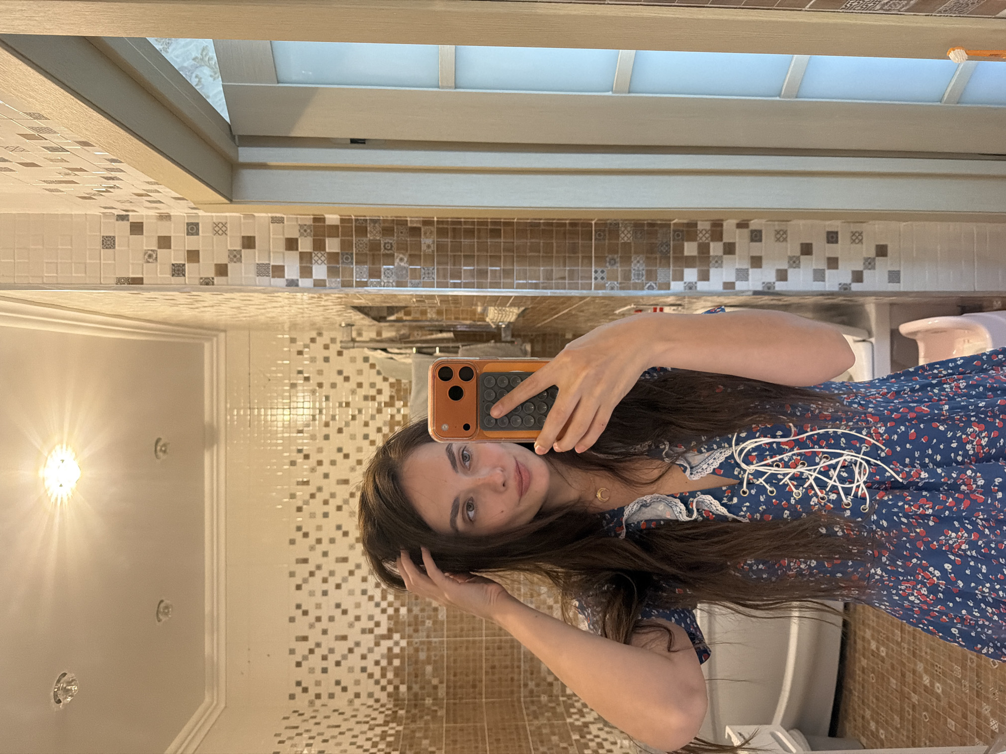The media file shows a young woman taking a mirror selfie in a bathroom. She is wearing a blue floral dress with white lace trim and white lacing detail at the neckline. She has long, dark brown hair and is looking at the camera with a slight smile. Her left hand is touching her hair, and her right hand is holding an orange-cased smartphone. The background is a tiled bathroom with beige and brown square tiles and a white ceiling. A light fixture on the left casts a bright glow. There is a glimpse of a toilet in the background. The image appears to be taken indoors, likely during the day given the lighting. The overall mood is casual and relaxed. There is no visible text in the image.
