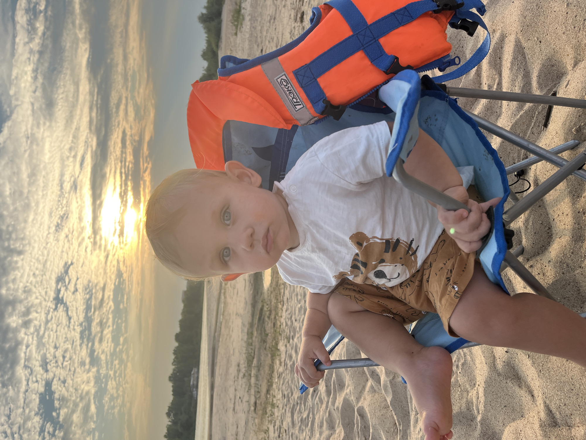 A young child with blonde hair and blue eyes sits in a blue folding chair on a sandy beach, with a life jacket draped over the back of the chair. The child wears a white t-shirt with a tiger graphic and brown shorts. In the background, a river or lake stretches towards a treeline, with a building visible in the distance. The sun is setting, casting a warm glow over the sky and water. The scene appears to be a waterfront area in Nizhny Novgorod, Russia.