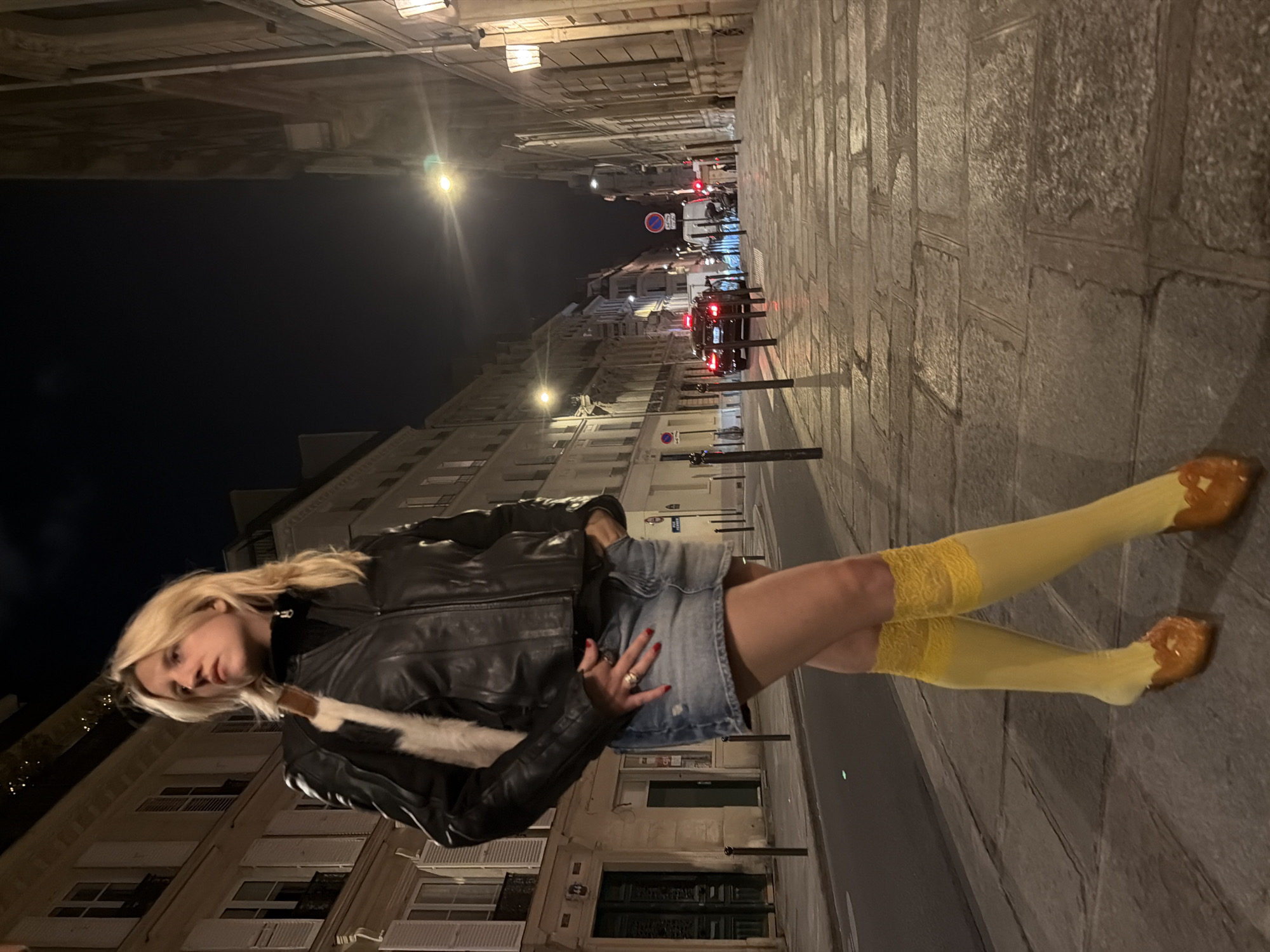 A full-body photograph captures a blonde woman posing on a street in Paris, France, at night. She stands on a stone-paved sidewalk, looking towards the camera with a neutral expression. She wears a black leather jacket, a denim mini-skirt, yellow thigh-high socks with delicate lace trim, and brown, possibly embellished, shoes. Her left hand, adorned with rings and red nail polish, rests on her skirt. A white fur or shearling detail is visible on her jacket's collar or lapel.
The urban scene is dimly lit by scattered streetlights and the warm glow emanating from windows of multi-story buildings that line both sides of the narrow thoroughfare. The architecture suggests a classic European city setting. In the background, a car with illuminated red taillights is visible on the street, along with several street signs, including a circular red and blue "no parking/stopping" sign. The sky is dark, confirming the nighttime setting. The ground consists of large, irregular stone pavers. No specific text is clearly legible on any signs.