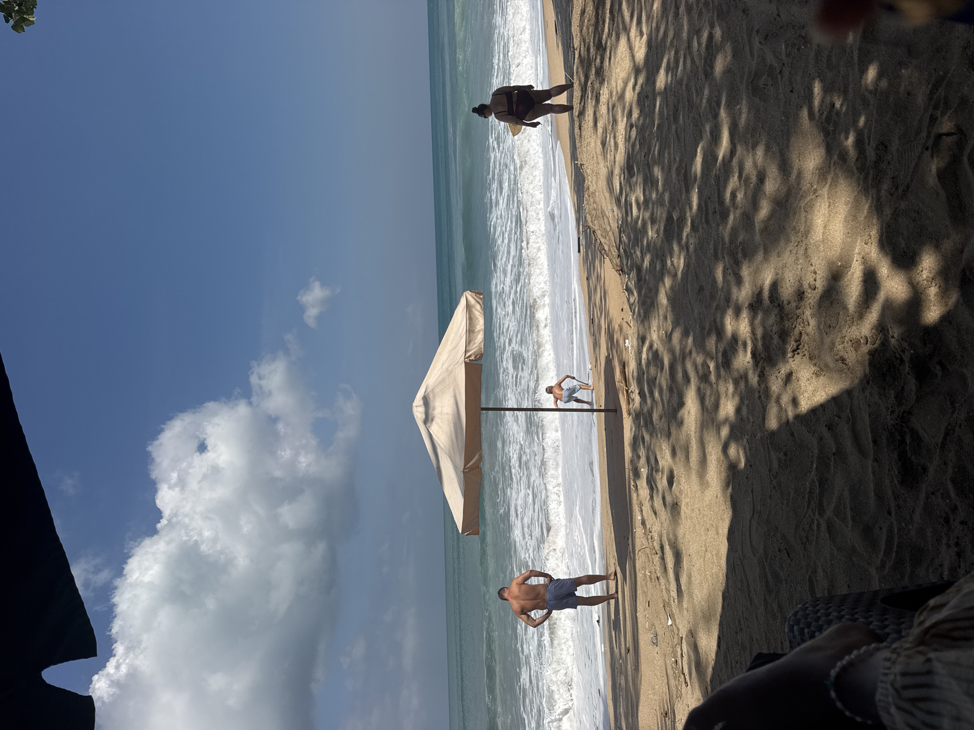 The image depicts a sunny day at Kuta Beach, Indonesia. The foreground shows sand with shadows of palm trees cast upon it, and a person's arm and leg are visible on the left, seated in a woven chair. In the mid-ground, a beach umbrella stands erected on the sandy shore. Further back, the ocean displays gentle waves and a turquoise hue under a clear blue sky with scattered white clouds.
Three individuals are present on the beach. A man, viewed from the back, stands near the water's edge wearing blue shorts. To his right, a young person is squatting and playing in the sand near the umbrella. On the right side of the frame, a woman with dark hair is standing, facing away from the camera, holding a surfboard. She is wearing a dark-colored bikini. The overall scene conveys a relaxed beach atmosphere.