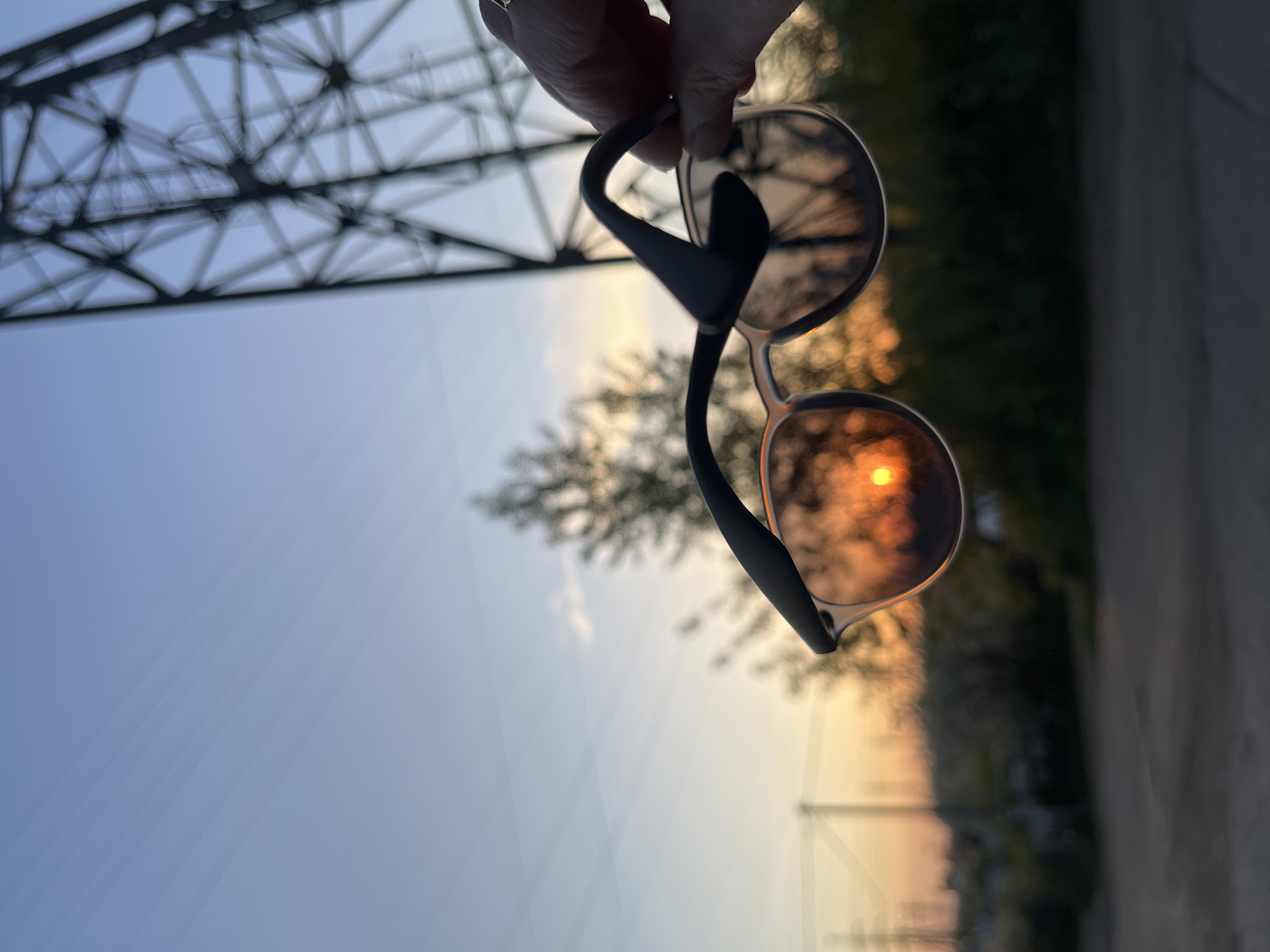 A hand holds a pair of sunglasses against the setting sun, with an electrical tower visible in the background. The location appears to be near a road in a residential area, potentially in the city of Chaikovsky, Russia. The image is taken from a low angle, looking up toward the sky. The sky is blue with streaks of orange and yellow where the sun is setting. The sun is seen through the bottom lens of the sunglasses, creating a glowing effect. The top lens shows the blurred reflection of the surroundings.