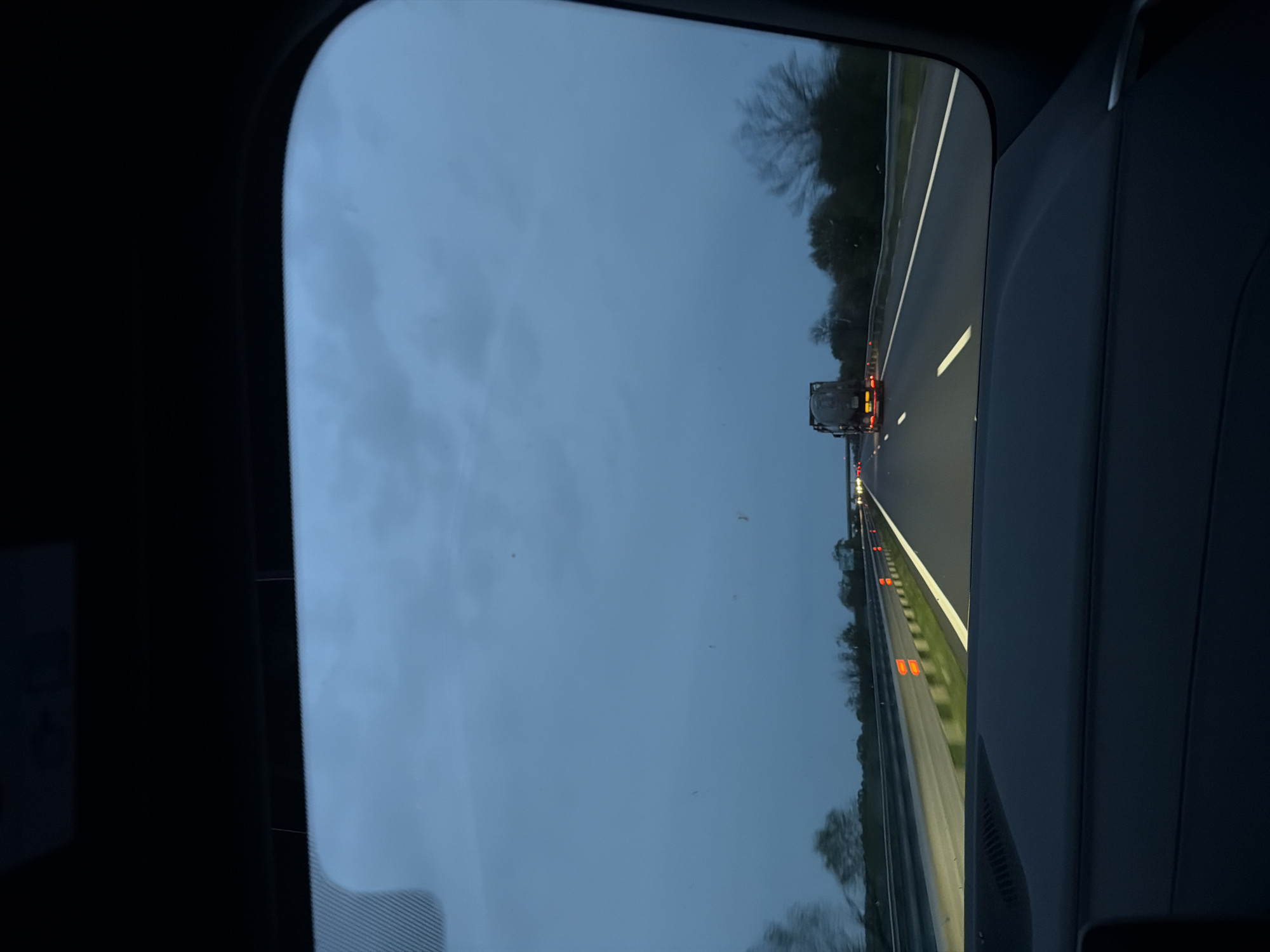The image depicts a view from inside a vehicle, looking out through a panoramic sunroof. The primary focus is on the highway ahead, with cars and a truck visible in the distance. Roadside reflectors line the shoulder of the road. The sky is overcast. There are trees lining the highway. The location is not Tsalapitsa, Bulgaria.