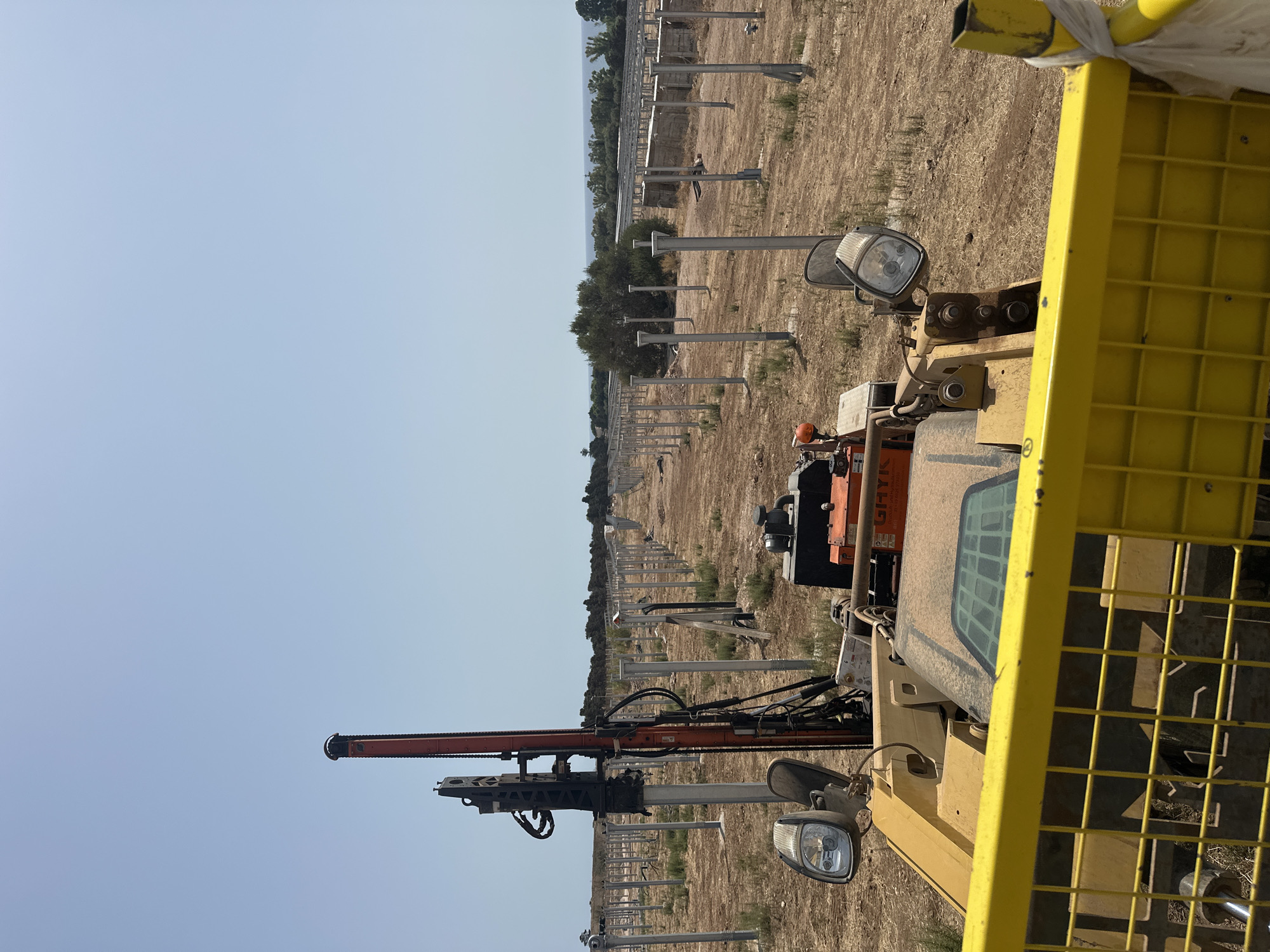 A bright, sunny day shows a construction site in Scicli, Italy, where metal poles are being driven into the ground, likely for a solar panel installation. The foreground is dominated by the yellow metal cage of a vehicle, with a portion of its dashboard, including a headlight, visible. Behind this, a large, orange and black drilling machine is positioned next to one of the metal poles, with hoses and mechanical parts extending from it. In the background, rows of similar metal poles stretch across a dry, dusty field, interspersed with sparse vegetation. Further back, trees and the distant sea are visible under a clear blue sky. There are no people visible in the image.