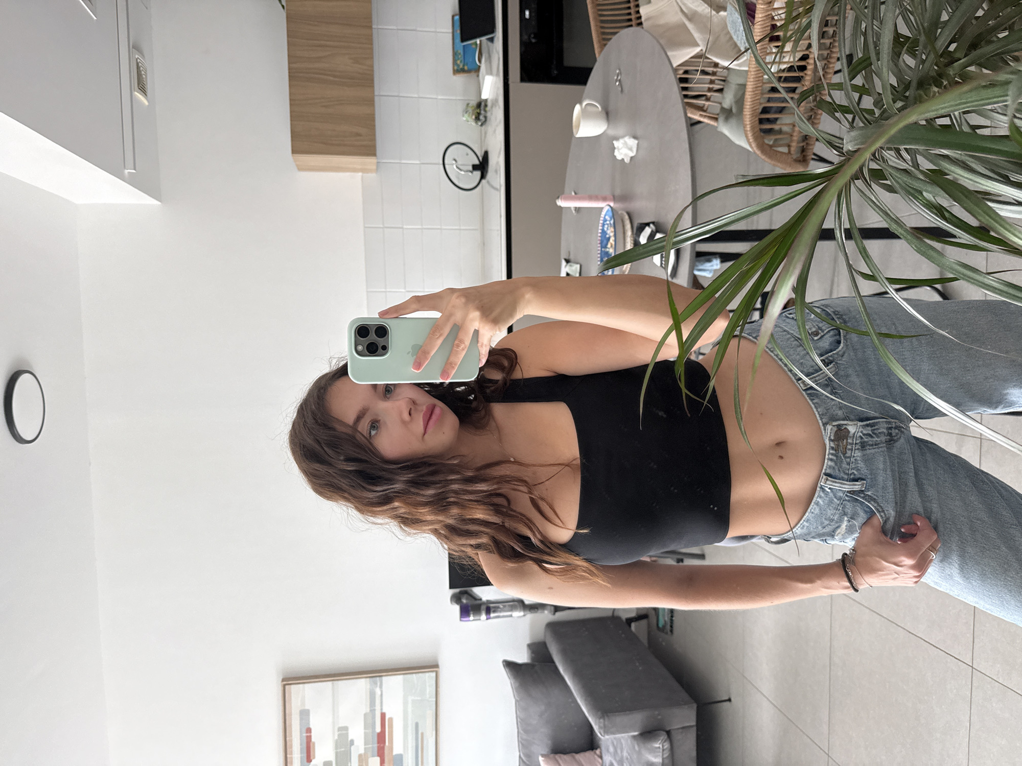A young woman is captured in a mirror selfie inside a modern apartment, likely in Ramat Gan, Israel. The image is oriented vertically, appearing rotated counter-clockwise. She has long, wavy brown hair and a neutral expression, holding a light green iPhone with multiple cameras to take the picture. She is wearing a black crop top that reveals her midriff and light blue, high-waisted denim jeans. Several rings adorn her right hand and a bracelet is on her left wrist.
The setting appears to be an open-plan living room and kitchen area with white walls and a light-colored floor. In the background reflection, a contemporary kitchen features white tiled backsplash and a wooden wall-mounted cabinet. A grey round dining table is visible with a white mug and a crumpled tissue on it, alongside a wicker chair. A large potted plant with long, thin leaves partially frames the shot on the right. Further back, a grey sofa with cushions and a stick vacuum cleaner are visible. A framed abstract artwork hangs on a wall, and a small black circular mirror is also present. The scene is brightly lit, suggesting it is daytime. There is no visible text in the image.
