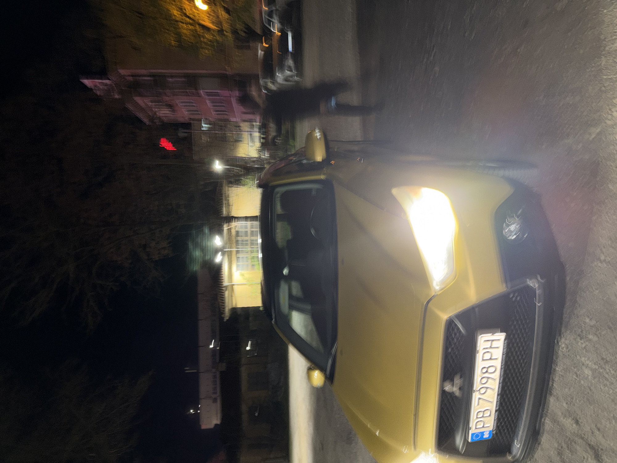 The image shows a gold Suzuki Swift parked on a street in Plovdiv, Bulgaria, at night. The car has a Bulgarian license plate "PB 7998 PH." The street is illuminated by streetlights. Buildings and trees are visible in the background. There is a blurred figure of a person walking next to the car.