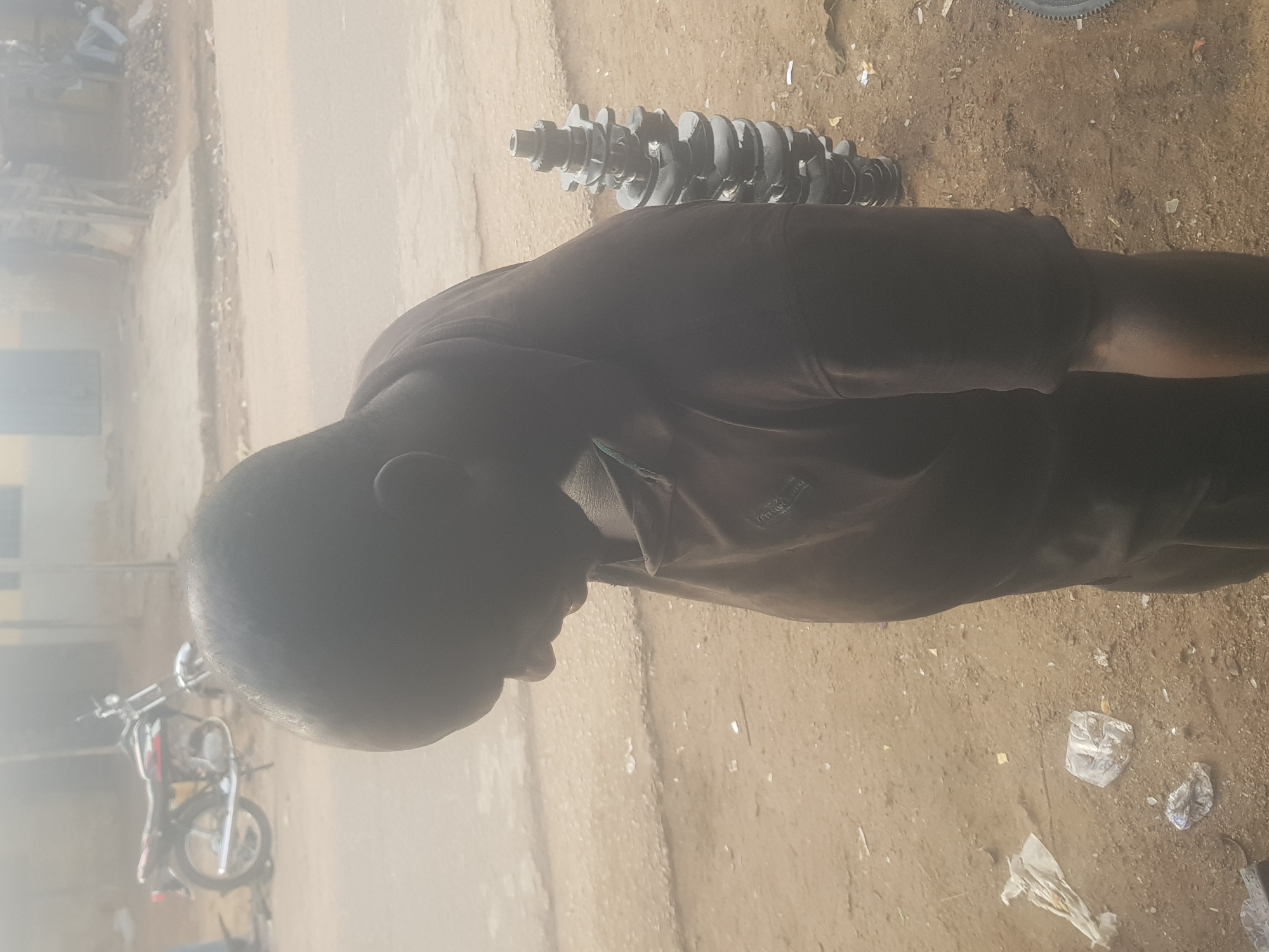 The image, rotated 90 degrees clockwise and set in Tukur Tukur, Nigeria, depicts an outdoor scene under bright daylight. In the immediate foreground, a person, viewed from the back and side, is the central subject. The individual appears to be male, with short dark hair, wearing a dark t-shirt, possibly black or dark brown, with a hint of green or blue fabric visible at the neck. The person is looking downwards and slightly to the left, seemingly at the ground or an object below.
To the right of the person, lying on the dusty, light brown ground, is a prominent metallic object resembling an engine crankshaft, dark grey in color. Various pieces of white plastic litter, such as crumpled bags, are scattered across the unpaved surface. In the blurred background to the left, a motorcycle is partially visible, suggesting a vehicle repair or parts sales context. Faint, indistinct outlines of buildings or structures can be seen in the far background.
The setting is an informal, unpaved outdoor area, possibly a street or a workspace for vehicle maintenance. The ground is dry and dusty. The lighting is strong and direct, characteristic of midday sun, casting some shadows. The person's posture suggests observation or contemplation rather than active work. A small, illegible logo or text, possibly "SPORT," is faintly discernible on the person's t-shirt. The overall scene conveys a functional, perhaps working, environment.