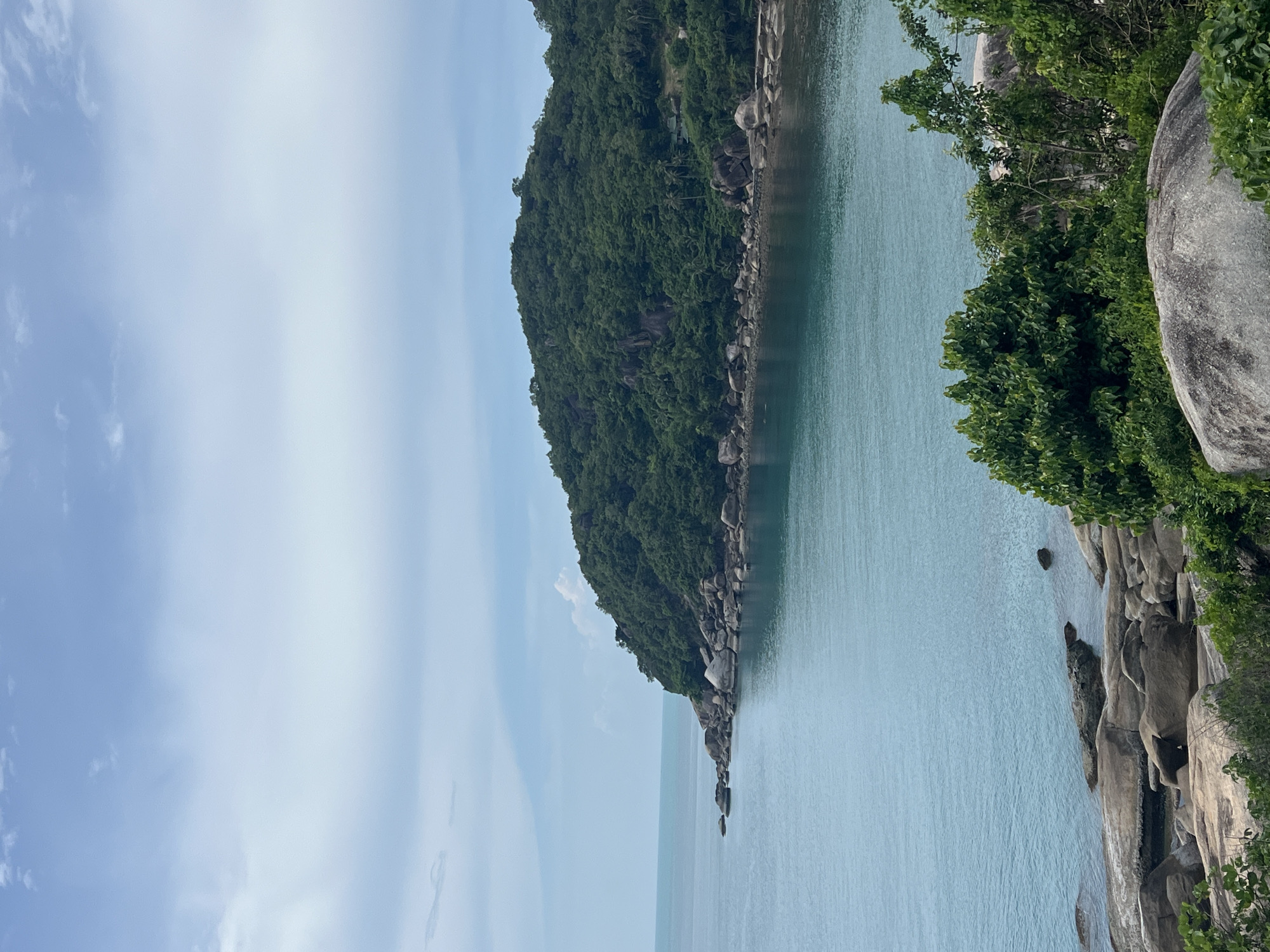 The image captures a scenic view of a tropical coastline. In the background, a lush green, tree-covered hill slopes down to meet the turquoise ocean. The shoreline is characterized by a rocky beach with large, smooth boulders. The water appears calm and clear, with gentle waves lapping against the rocks. The sky above is a pale blue, with scattered white clouds. The overall impression is one of natural beauty and tranquility, consistent with the tropical environment of Baan Chaweng Noi, Thailand. There are no people or animals visible in the image.