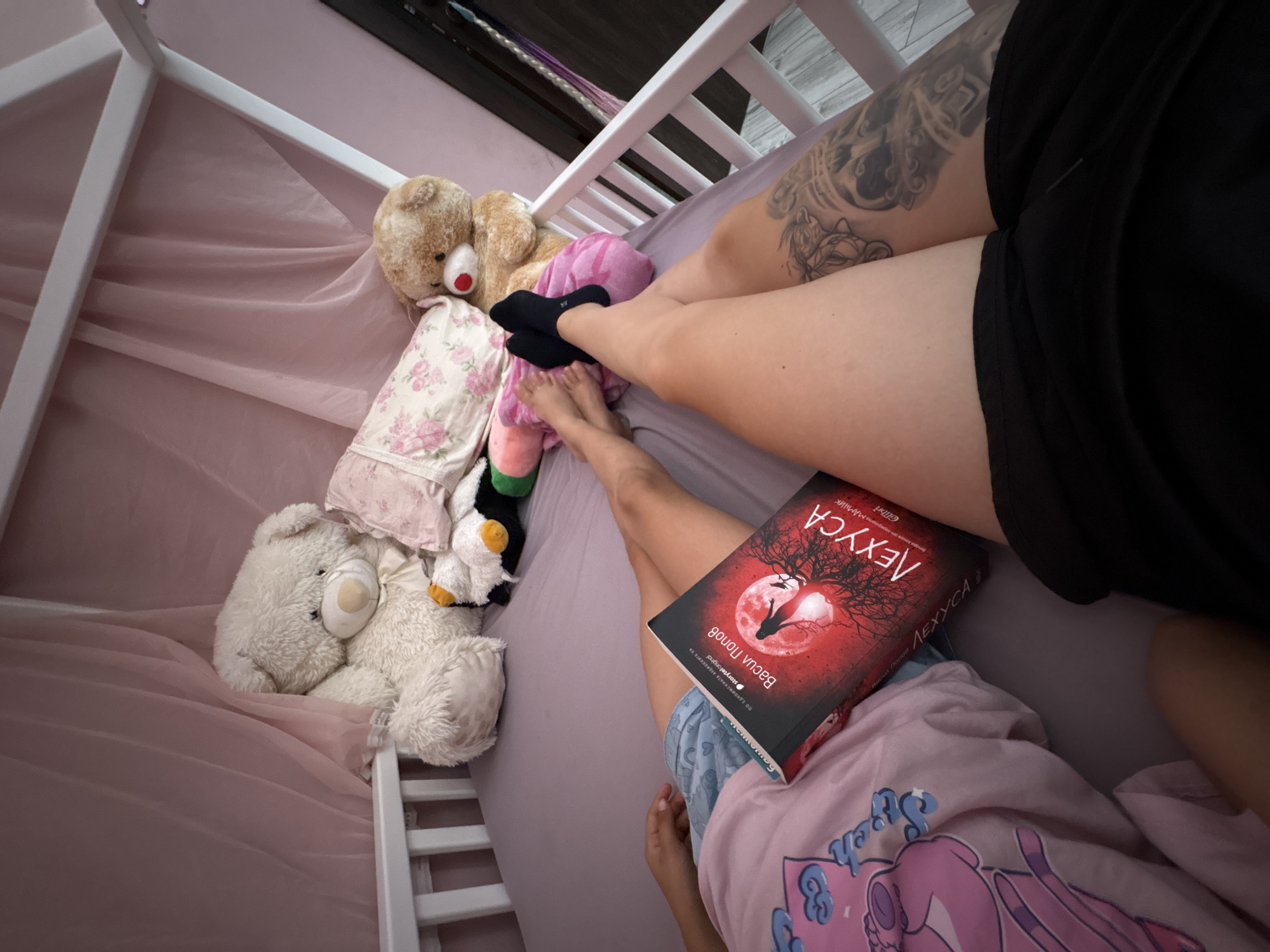 The image shows a person lying in a crib-like bed, which is decorated with pink sheer fabric. The bed contains several stuffed animals, including a large white teddy bear and a smaller brown teddy bear. A book titled "ЛЕХУСА" is open on the bed, with its pages resting on the person's legs. The person is wearing shorts and black socks, and one leg displays a prominent tattoo. The environment appears to be a bedroom, likely a child's room, given the presence of the crib and stuffed animals. There is no information in the image that would indicate a specific location within the city of Oreshak, Bulgaria.