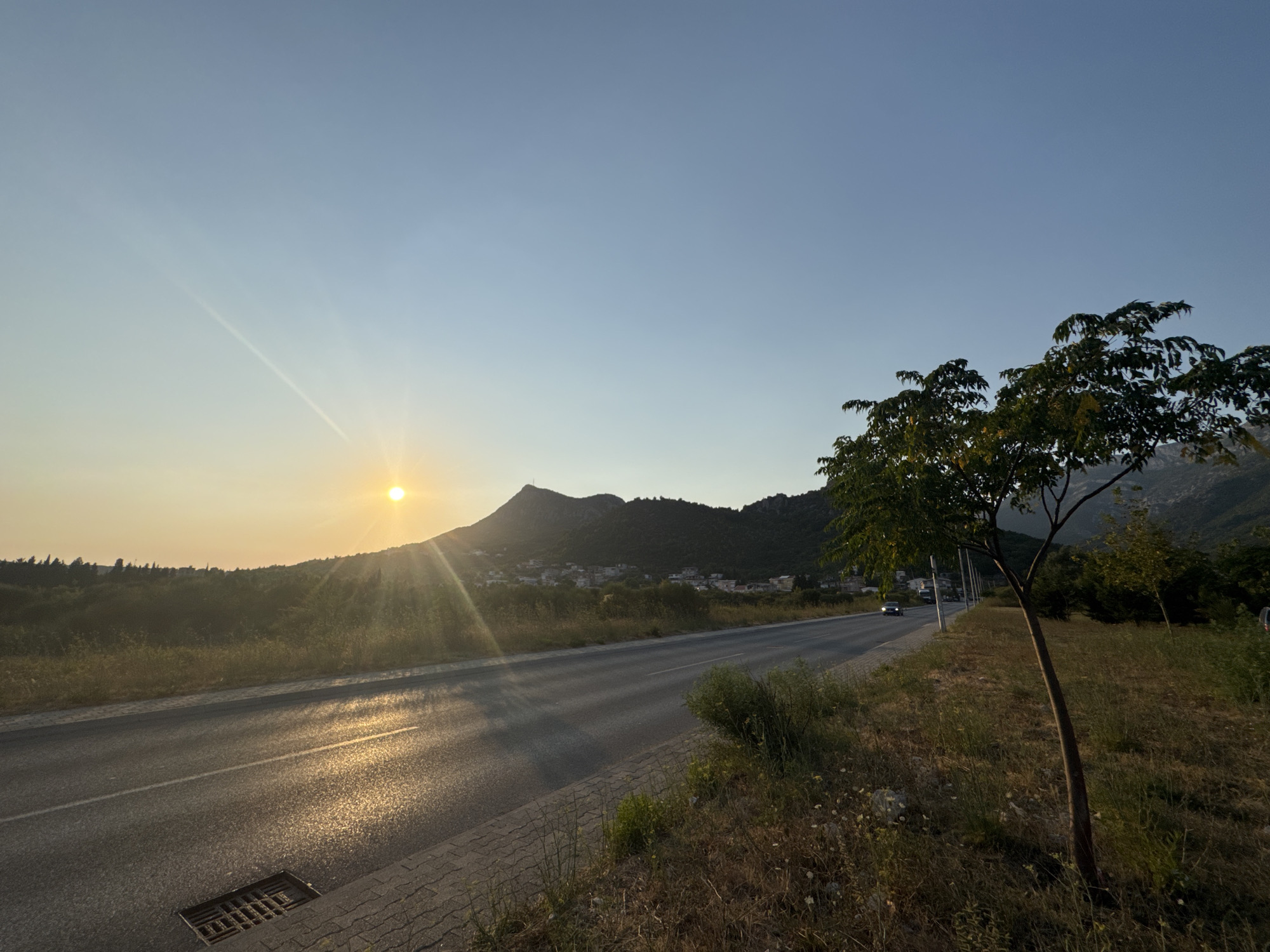 This is a horizontal, eye-level, outdoor photograph taken at sunset in Sutomore, Montenegro. The sun is low in the sky, casting a warm, golden light across the scene. A paved road runs diagonally from the lower left to the middle right of the frame, with a white line marking the center. The road is wet and reflects the sunlight. To the left of the road, there is a patch of dry grass and vegetation. A metal grate is visible on the edge of the road in the lower left corner.
In the background, a mountainous landscape is visible, covered with trees and dotted with buildings of a small town. A single tree with green leaves stands prominently in the right foreground, its trunk slender and dark. The sky is a clear, pale blue, with a faint contrail visible in the upper left quadrant. The overall impression is one of a peaceful, scenic road at dusk.