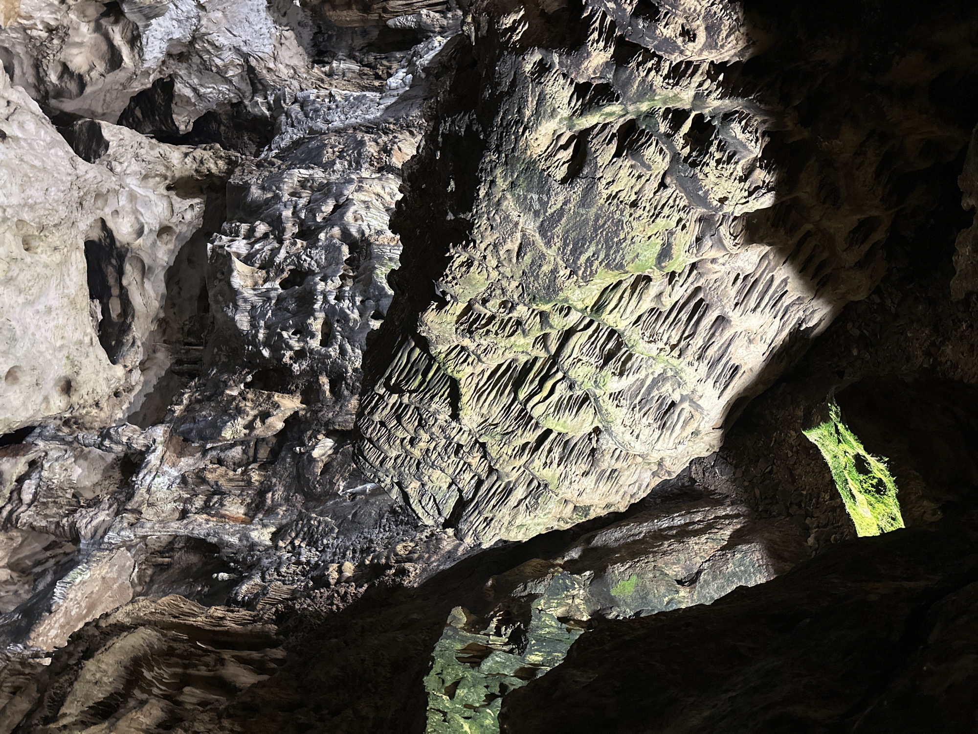 The image shows the interior of the Magurata Cave in Raianovtsi, Bulgaria. The cave walls are composed of light gray and dark brown rock formations with varying textures, including stalactites and porous surfaces. Some areas have a green tint, possibly due to moss or algae. Light enters the cave through openings in the rock, creating areas of illumination and shadow. No people are visible in the image.
