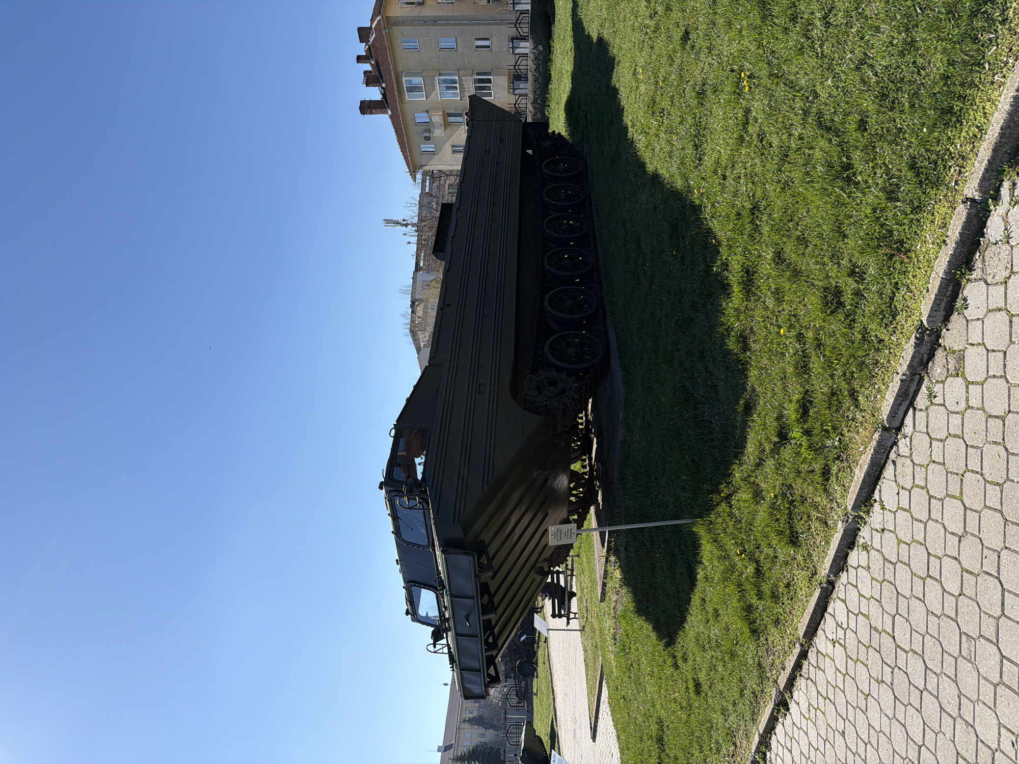 A sideways shot shows a tracked vehicle on display in a grassy area adjacent to a stone-paved walkway. The vehicle is dark green and appears to be an amphibious transport or combat vehicle, with visible tracks and a flat, boxy shape. It is positioned between the lawn and the walkway, with the edge of the lawn bordered by a concrete curb. Buildings with windows are visible in the background. The sky is clear and blue. The scene appears to be in a city park or museum setting.