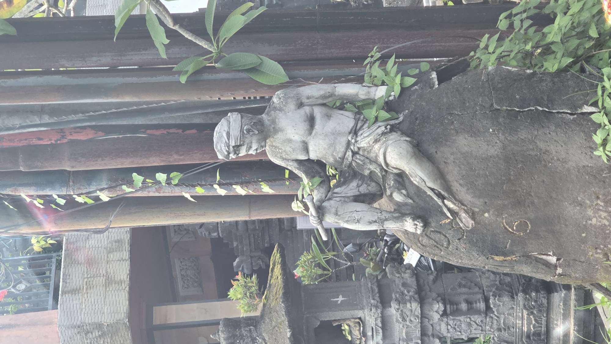 The image displays a weathered stone statue situated outdoors, likely within a garden or temple area in Ubud District, Indonesia. The central subject is a grey, masculine figure carved from stone, depicted in a kneeling or crouching position. The statue features a muscular physique, with a cloth covering its eyes like a blindfold and a loincloth around its waist. The figure holds a short, staff-like object pointing downwards with both hands.
The statue shows significant signs of age and weathering, including noticeable cracks on its legs and the large, dark grey stone slab it rests upon. Green vines and leafy plants are integrated into the scene, growing around and on parts of the statue and its base. Larger green leaves are visible in the upper sections of the frame.
In the background, there are horizontal, weathered structures in shades of brown, red, and dark grey, possibly wooden beams or parts of an old building. Behind the statue and to its right, intricate dark stone carvings, typical of traditional Balinese architecture, are visible, suggesting a temple or similar cultural structure. Further back, a textured grey wall can be seen. The scene is illuminated by natural daylight, indicated by a lens flare in the lower left, signifying direct sunlight. On the stone base near the statue's leg, faint, indistinct symbols or markings are present.