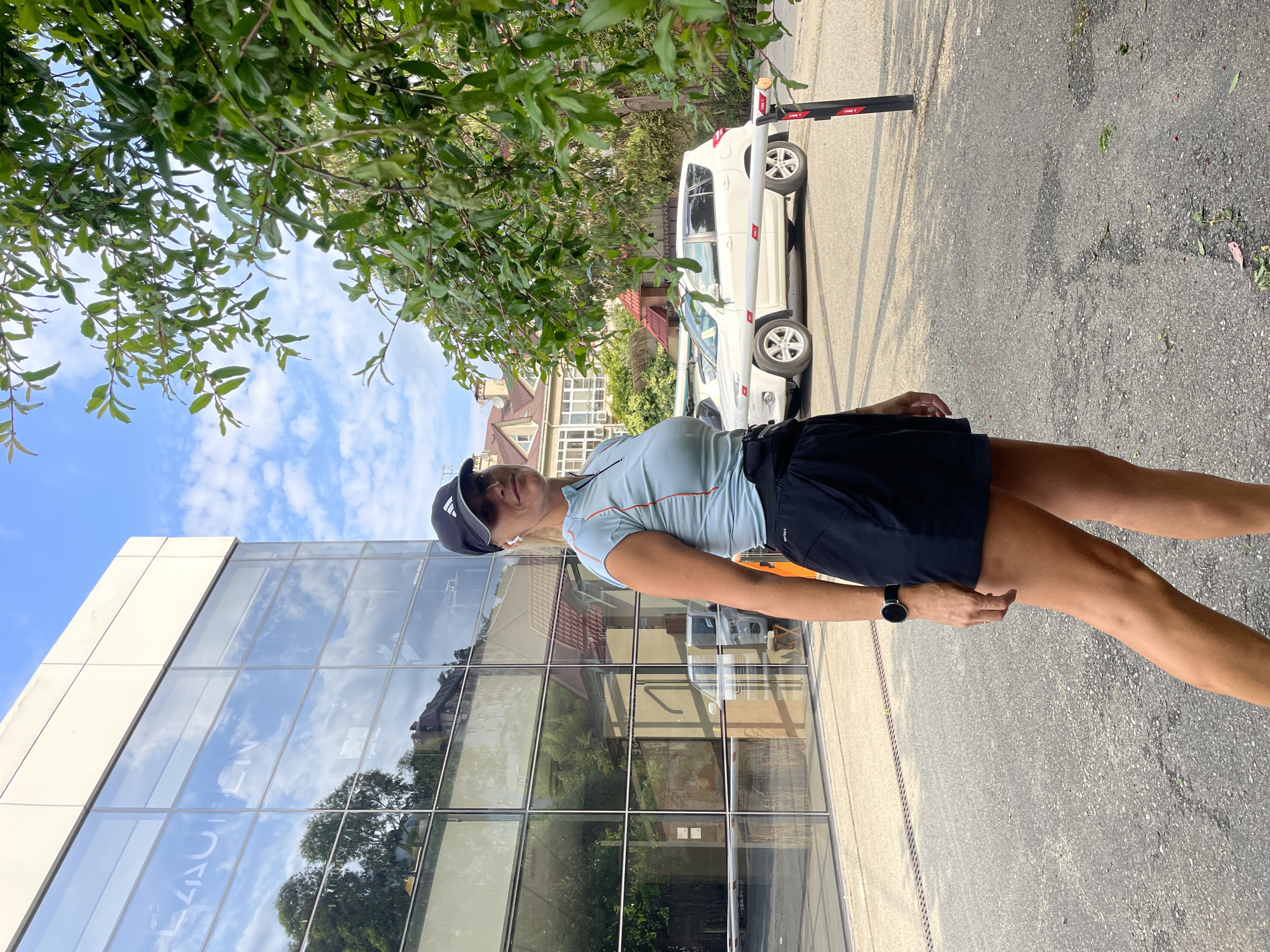 A woman wearing a blue athletic shirt, black shorts, and a black baseball cap stands outdoors. She has a smartwatch on her left wrist. In the background, a white SUV is parked in front of a modern glass building. Trees with green leaves are visible on the right side of the image. The scene appears to be in the city of Sochi, Russia.