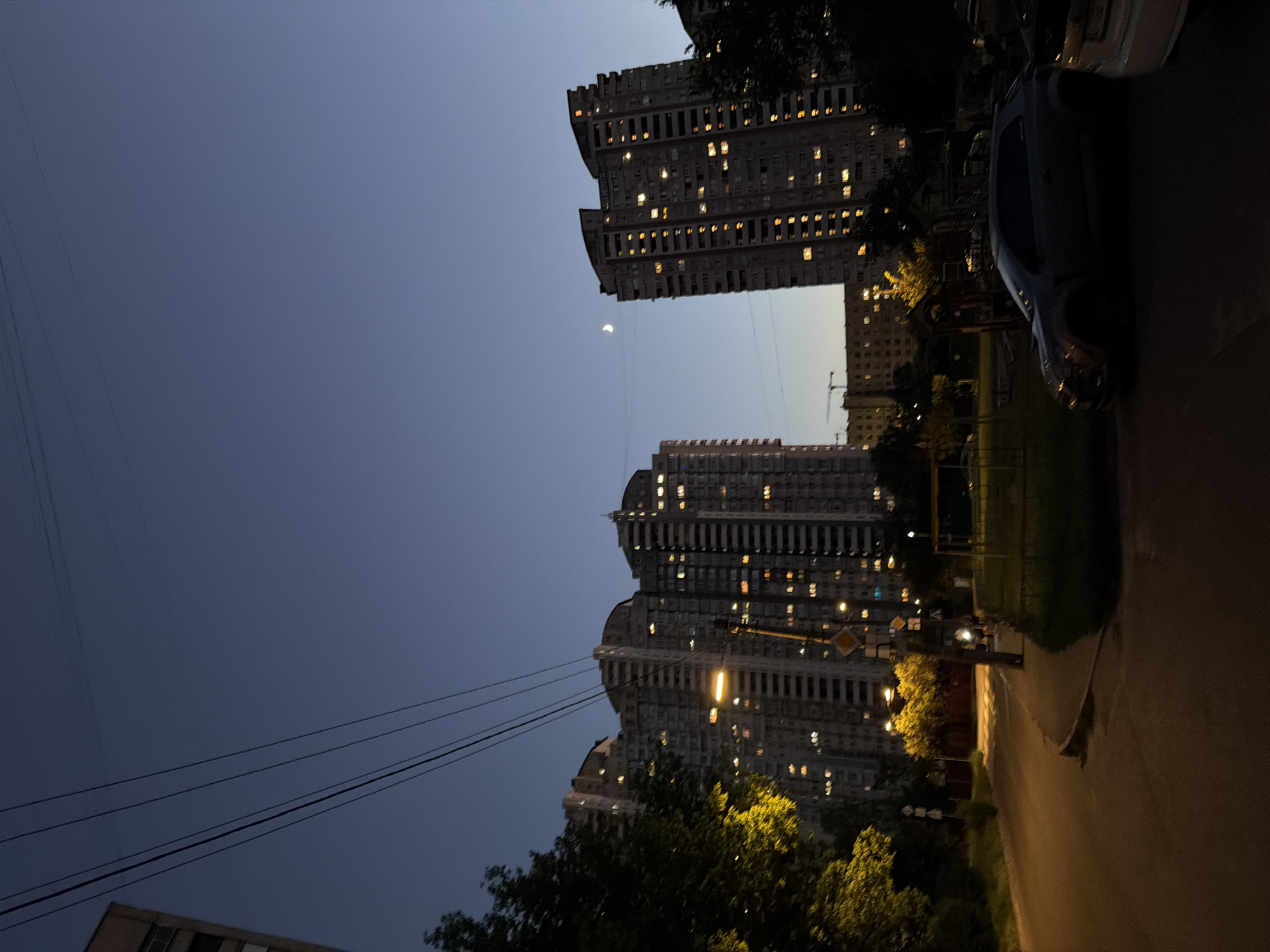 The image depicts a twilight scene in Kyiv, Ukraine, with two tall, multi-story apartment buildings dominating the background. The buildings are illuminated by numerous yellow-lit windows, suggesting occupancy. A crescent moon is visible in the dark blue sky above the right-hand building. In the foreground, a paved road curves to the left, bordered by a sidewalk and a grassy area. A dark-colored hatchback car is parked on the right side of the road, with another vehicle partially visible next to it. Trees with bright green foliage are present on the left side of the road, and a streetlamp casts a warm glow on the asphalt. Several utility poles and wires crisscross the scene. The overall atmosphere is calm and residential, characteristic of a city neighborhood at dusk.