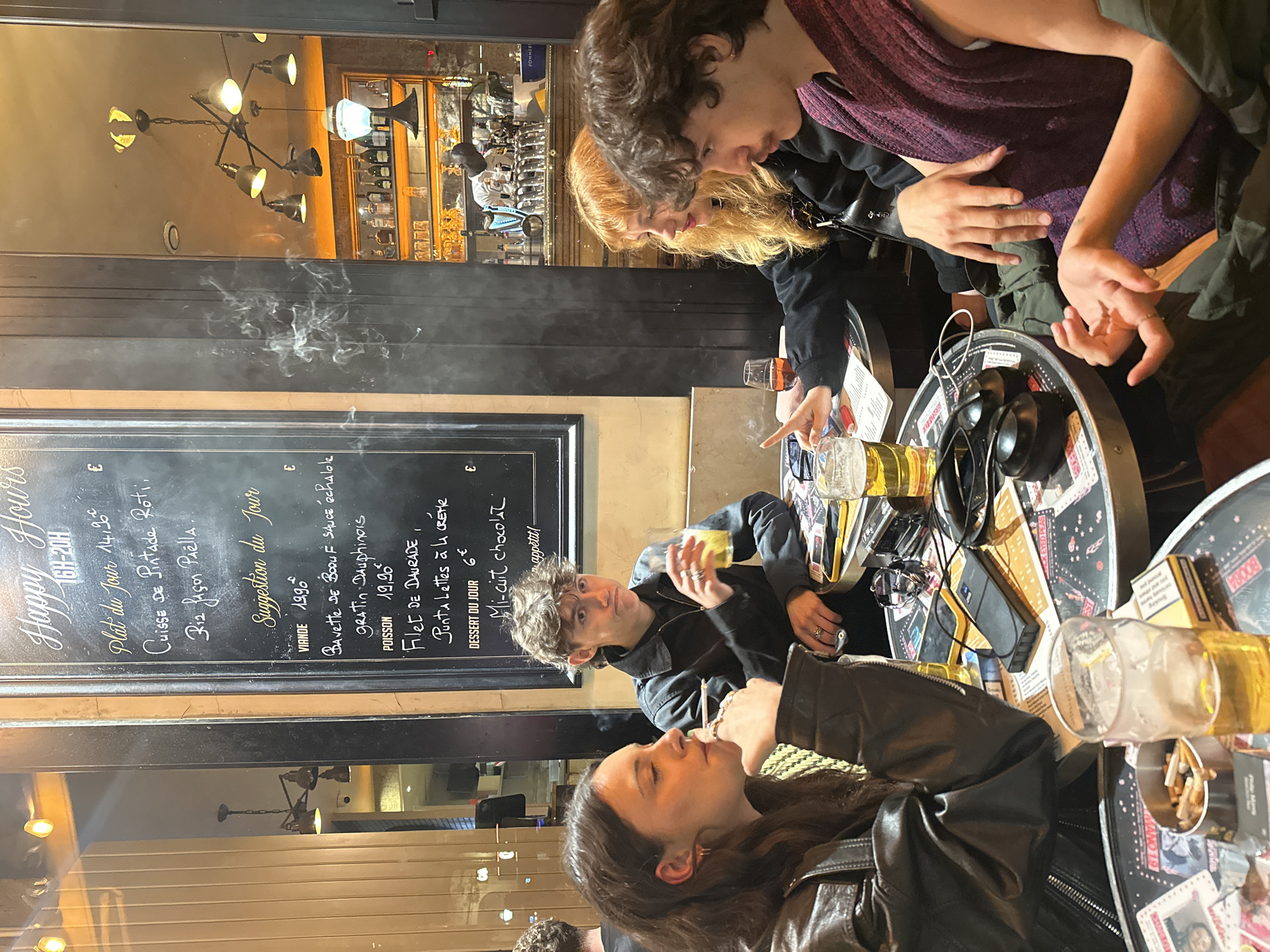 The image depicts a group of people sitting at tables outside a bistro, likely in Paris, France. The scene features a chalkboard menu in the background, displaying "Happy Hours" and "Suggestion du Jour," as well as French dishes and prices.
Several individuals are seated at the tables, engaged in conversation. One person is smoking, with smoke visible above the table. The tables are cluttered with beer glasses, ashtrays with cigarette butts, and personal items like headphones. The bistro's interior is reflected in the glass behind the chalkboard menu, showing light fixtures and shelves with bottles. The atmosphere suggests a casual, social gathering at a Parisian cafe.