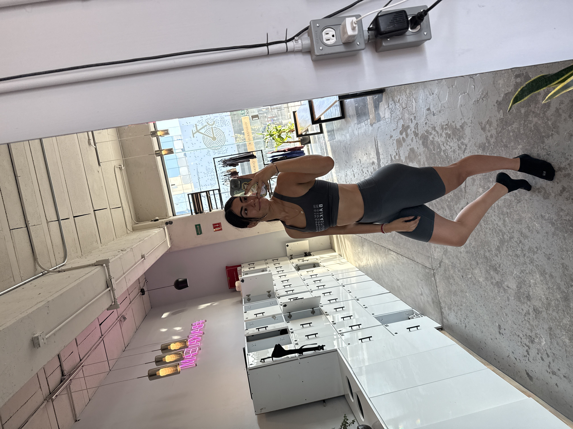 A young woman takes a mirror selfie in what appears to be a modern fitness or cycling studio. The image is captured from her perspective, showing her reflection in a large mirror that forms one wall of the room. She is standing with her left leg bent, holding a smartphone with a patterned case up to capture her image. She has dark hair, is wearing a dark grey sports bra with the text "BAIKAS" (reversed in the reflection) and matching athletic shorts, along with black socks. She has a subtle smile on her face.
The setting features a bright, industrial aesthetic with a concrete-textured floor and white walls and ceilings. To the woman's right, a long bank of white lockers with black handles extends into the distance; some locker doors are open, revealing their interior, and the number "19" is visible on one. Above the lockers, a pink neon sign spells out "BIKAS". Hanging light fixtures with cylindrical shades are also visible.
Behind the woman, a large window reveals an urban exterior with other buildings, some foliage, and bright daylight. A white decal on the window displays a stylized bicycle logo and the text "BIKASSTUDIO", clearly indicating the nature of the establishment. Electrical outlets and conduit are visible on the upper wall, and a green emergency exit sign is also present. The overall atmosphere suggests a clean, contemporary gym or workout space.