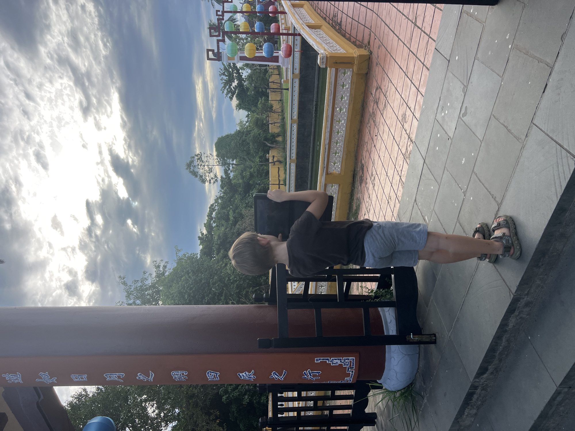 A young boy stands on a tiled pathway in front of a maroon pillar with white Chinese characters inscribed vertically. He is holding a dark-colored tablet in his hands, pointed towards the right side of the frame. The boy is wearing a dark t-shirt and light grey shorts, along with sandals. To his right, a yellow tiled area leads to a body of water, with a decorative white railing along its edge. Beyond the water, there is a white building with decorative red trim and a row of colorful lanterns hanging from poles. The background consists of lush green trees and a cloudy sky. The scene appears to be located within the grounds of the Imperial City in Hue, Vietnam, given the architectural style and the presence of Chinese characters.