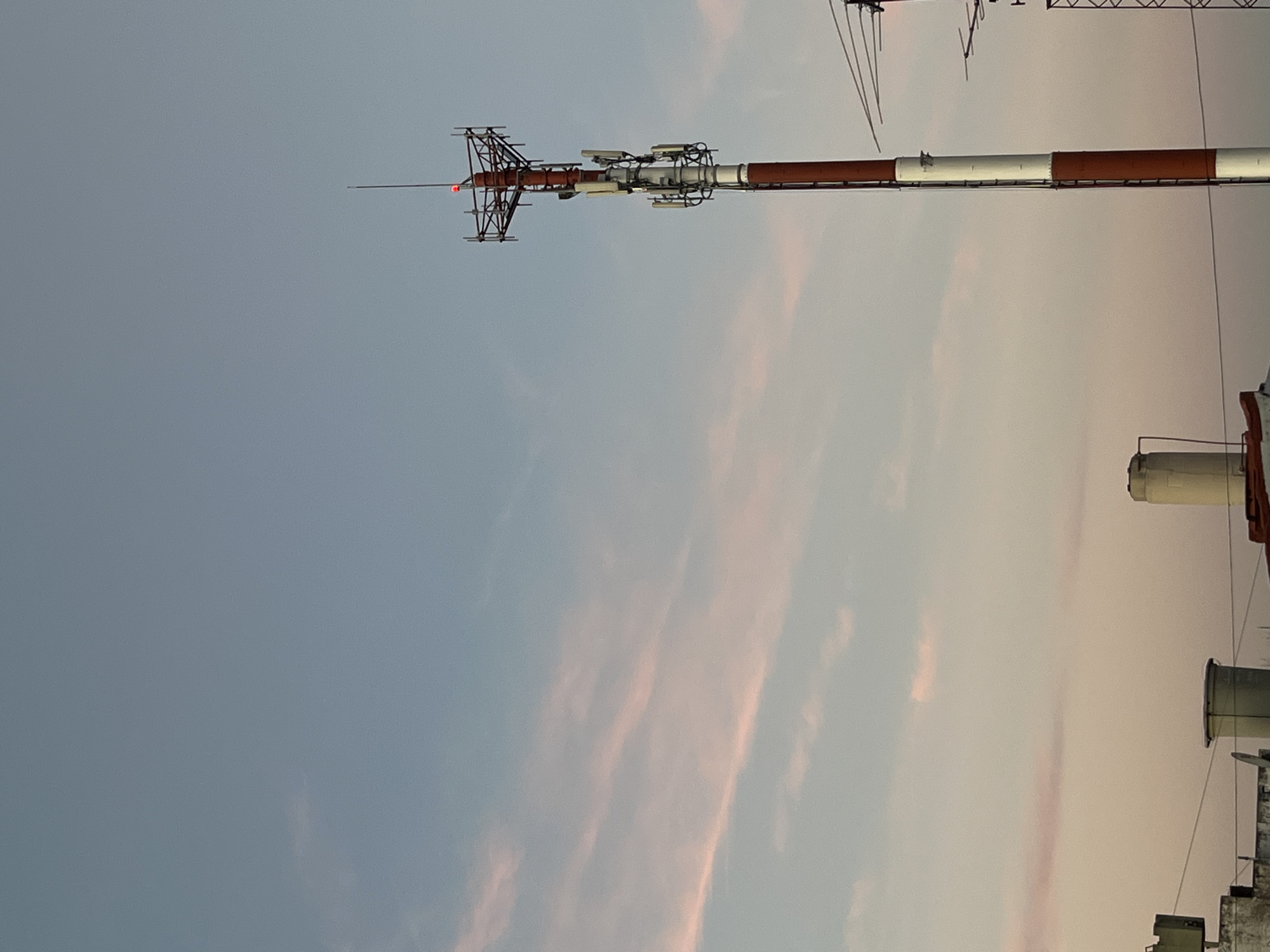The image displays a tall, slender communication or antenna tower set against a vast, evolving sky, located in Buenos Aires, Argentina. The tower, a prominent feature on the right side of the frame, stands vertically with distinctive horizontal bands of reddish-brown and white paint. At its very top, a complex metallic structure, likely an antenna array or a lightning rod, is visible, featuring a small, glowing red light. Below this, various smaller antennae and equipment boxes are attached to the main mast.
The sky occupies the majority of the background, exhibiting a soft color gradient. It transitions from a lighter, somewhat grayish-blue on the left to warmer, delicate pink and orange hues on the right, characteristic of either sunrise or sunset. Sparse, wispy clouds are subtly colored by the low sun, indicating clear weather. In the bottom right corner, parts of a building's rooftop are visible, including what appear to be water tanks or ventilation units, suggesting an urban environment. No specific activity or event is taking place, and there is no visible text in the image. The scene conveys a sense of calm observation of urban infrastructure against a picturesque sky.