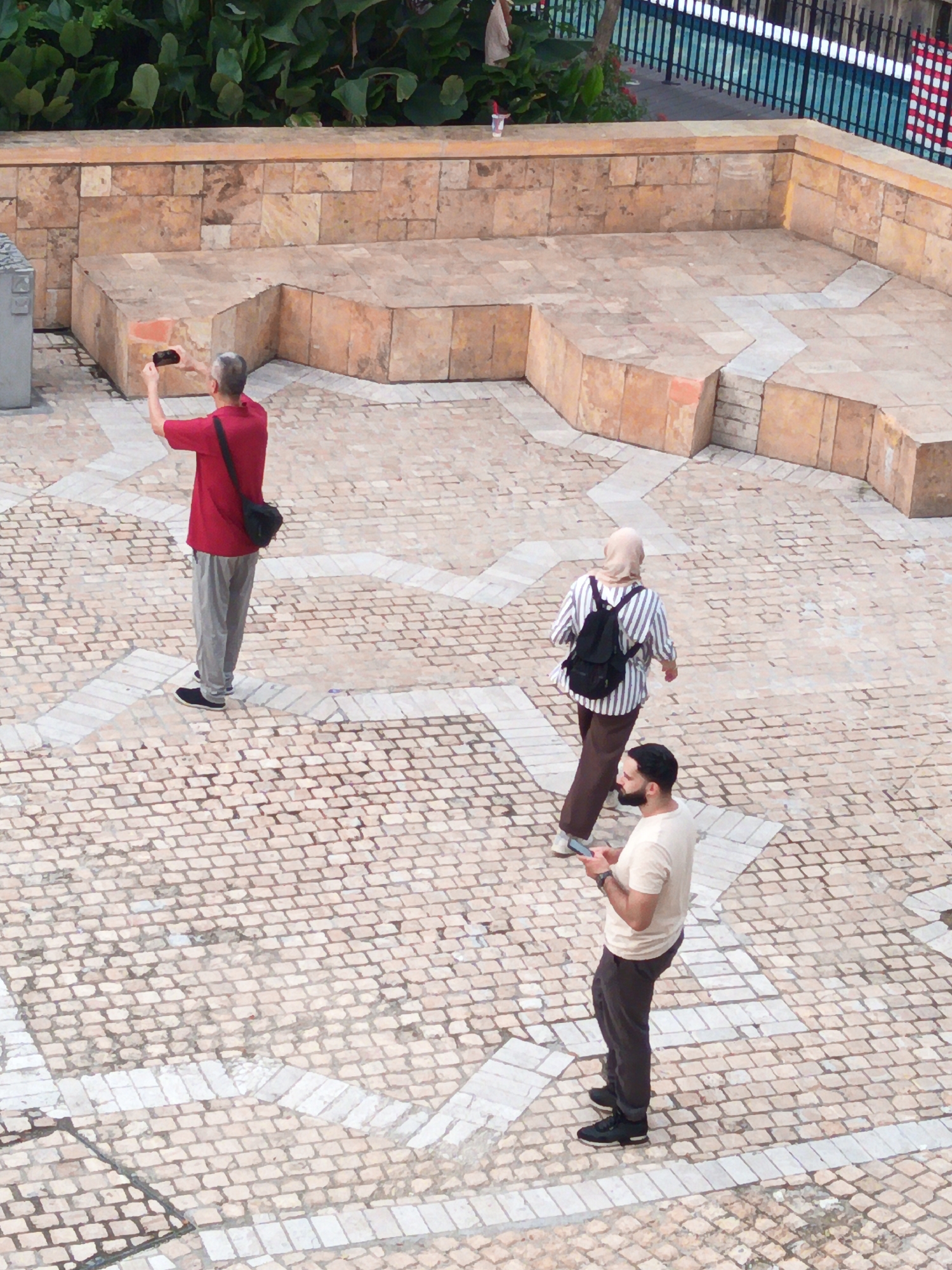 An aerial view captures three individuals on a cobblestone plaza in Kuala Lumpur, Malaysia. The plaza features a raised, tiered seating area constructed from large, light-brown stones. Behind this, a lush green foliage backdrop is partially visible. To the right, a black metal fence with blue and white elements suggests a border or pathway.
In the foreground, a man in a red t-shirt and grey pants stands facing away from the camera, holding a black smartphone at arm's length, appearing to take a photo. A black messenger bag is slung across his right shoulder. To his left, a woman wearing a light-colored hijab, a striped long-sleeved shirt, and dark brown pants walks away, carrying a black backpack. To the right of the woman, a man with dark hair and a beard, wearing a light-colored t-shirt and dark pants, is looking down at his black smartphone, holding it in his right hand.