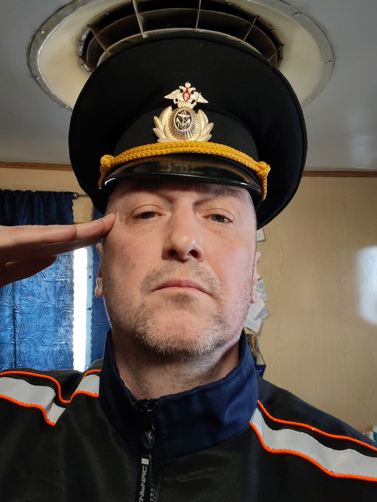 A medium eye-level indoor shot captures a man in uniform saluting. The man, wearing a black peaked cap adorned with a golden emblem and a gold braided trim, has a stern expression. He's also wearing a black jacket with a navy blue collar and orange and grey reflective stripes. His left hand is raised in a salute, obscuring part of the left side of his face.
Above his head is a circular ceiling fixture with a white rim and a dark interior, suggesting ventilation or lighting. The background includes a blue curtain on the left and a beige wall on the right, with some papers attached to it. The location, Rybinsk, Russia, is mentioned for context but is not visually confirmed in the image. The man's actions and attire suggest a military or official capacity.