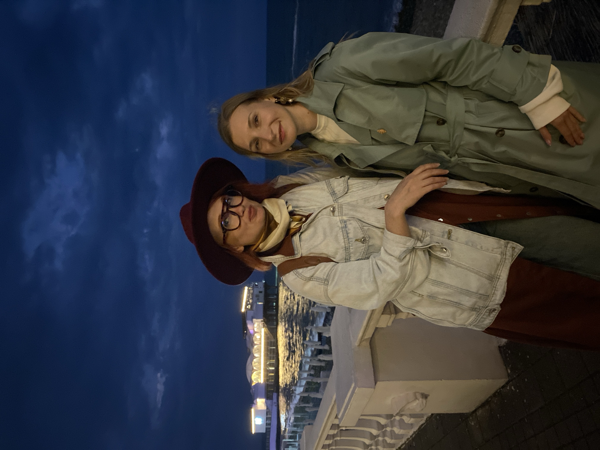 Two women are standing next to each other on a balcony with a view of the Sochi pier in Russia at night. The woman on the left is wearing a maroon hat and glasses, a denim jacket, and a scarf. The woman on the right is wearing a light green trench coat and gold earrings. The Sochi pier is illuminated with lights. The sky is dark blue.