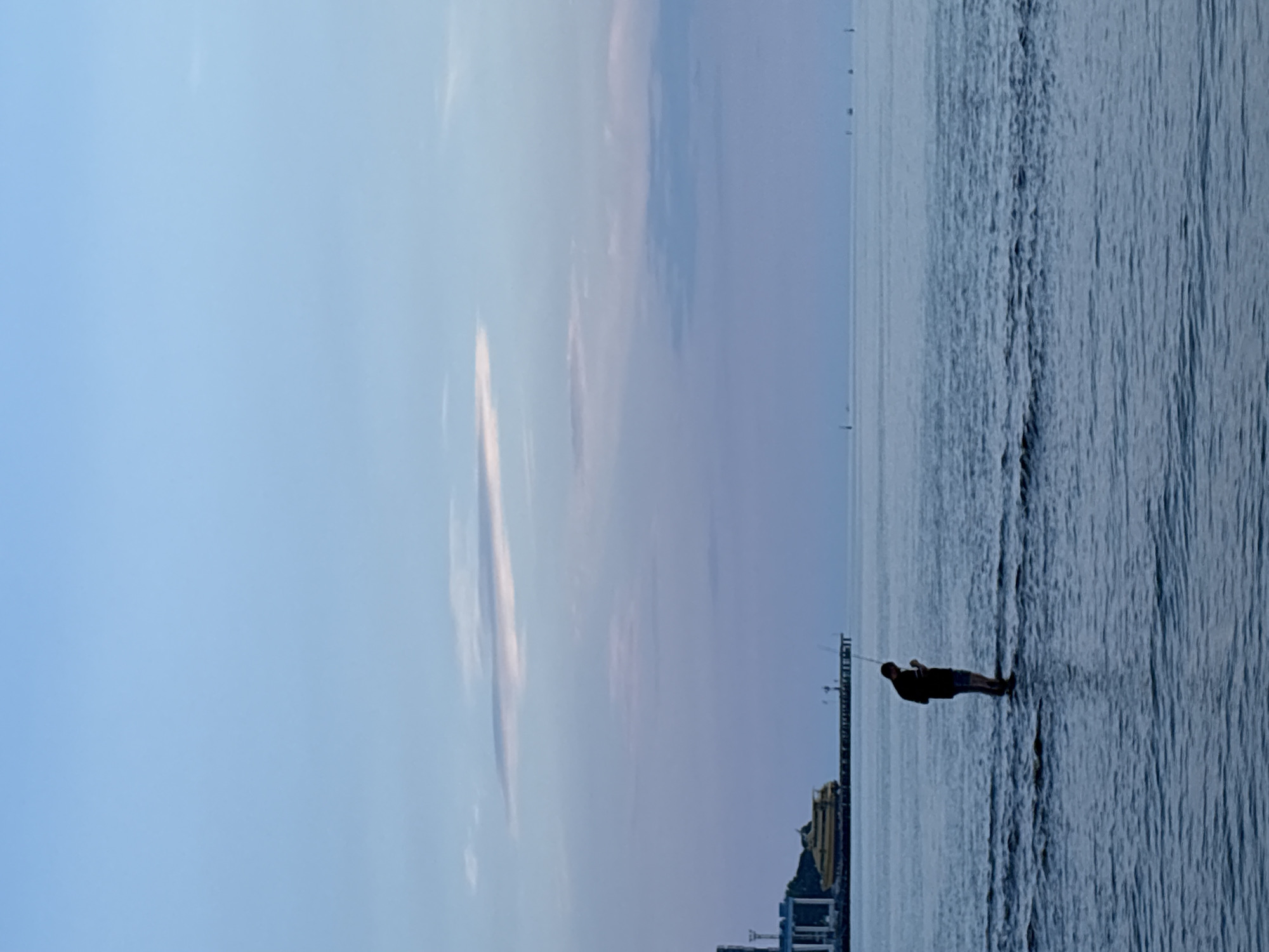 The image depicts a serene evening scene at a beach in Anapa, Russia. The sky is a gradient of soft blues and purples, with a few wispy clouds. The calm sea reflects the twilight colors. In the foreground, a lone figure, silhouetted against the water, stands in the shallows, seemingly engaged with something in their hands. In the middle ground, a long pier stretches out into the sea, with a yellow vessel docked at its end. Further in the distance, several small boats are visible on the horizon, adding to the peaceful atmosphere. The overall impression is one of tranquility and solitude.