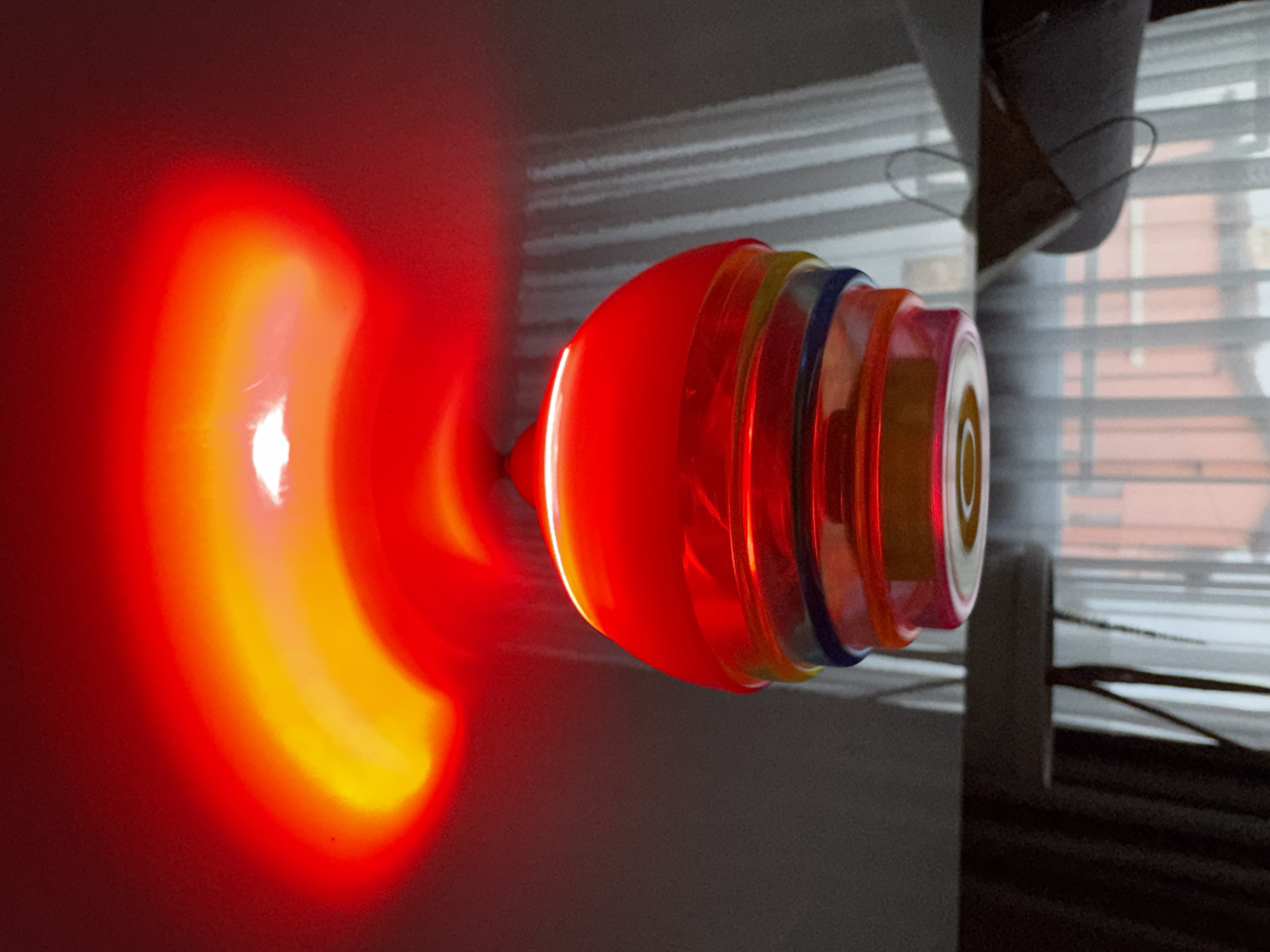 This media file captures a close-up indoor scene dominated by a brightly glowing, translucent object. The object, possibly a spinning top or a decorative lamp, is oriented vertically on the left side of the frame, emitting intense red and yellow-orange light. Its core is a bright, almost white-yellow, which gradually radiates outwards into vivid orange and then deep red, illuminating the immediate dark surroundings. The object itself appears to be constructed from layered, translucent materials, with distinct rings of red, orange, yellow, and a darker blue near its right side.
In the background, on the right, a window with horizontal blinds is visible, allowing soft, diffused light to enter the space. Through the blinds, vague shapes suggestive of buildings or structures can be faintly discerned in the distance. A dark, possibly fabric object, resembling a curtain or piece of clothing, hangs in the upper right portion of the frame. The overall lighting suggests a dimly lit room, possibly at dusk or night, highlighting the intense luminescence of the primary object. No people or visible text are present in the image. While the location context of Varna, Bulgaria, is provided, there are no specific visual cues within the image to identify this city.