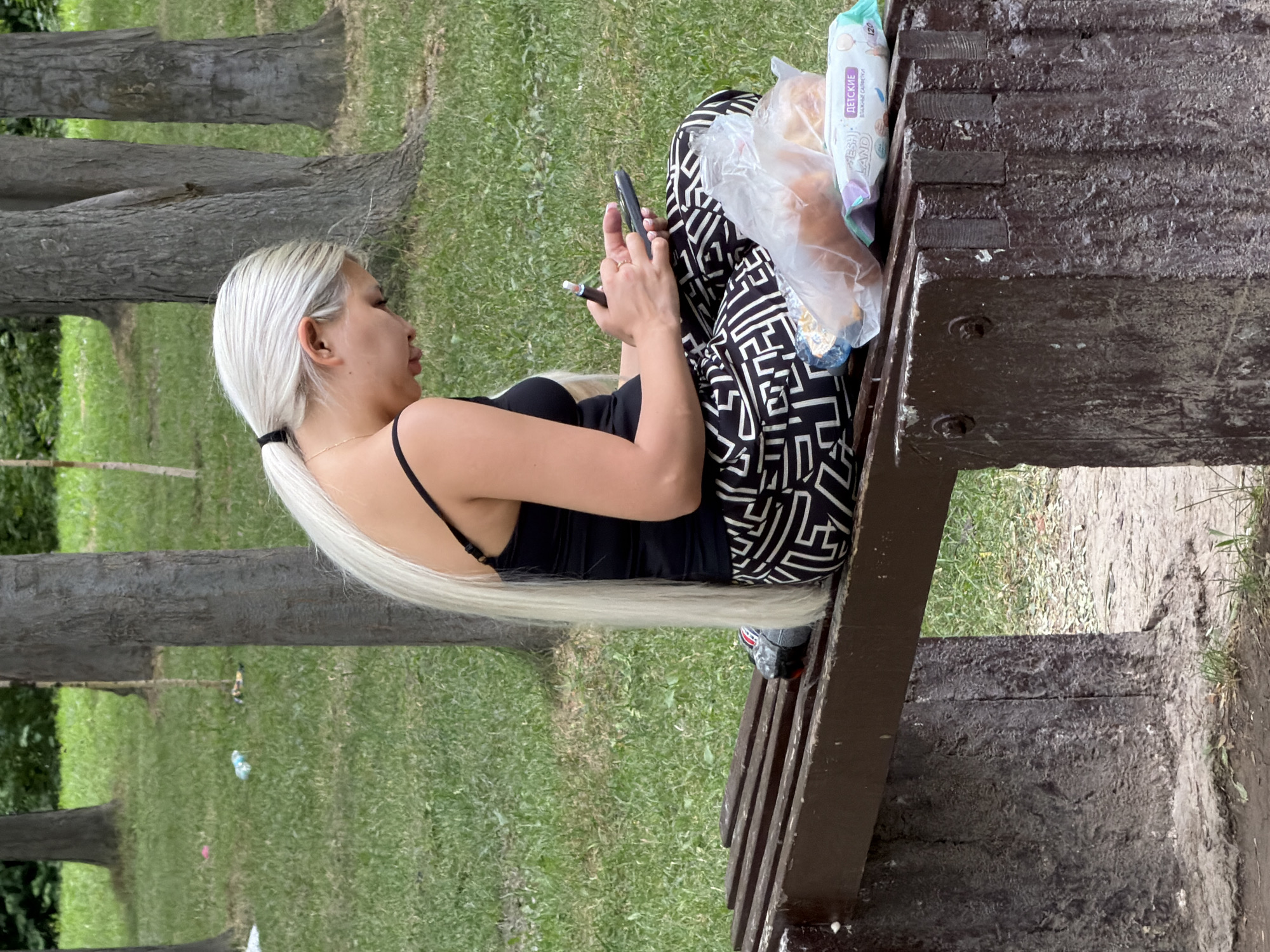 A woman with long blonde hair pulled back in a ponytail sits on a wooden park bench. She wears a black tank top and black and white patterned pants. The woman is holding a smartphone and a vape pen. To her right on the bench are a plastic bag containing food items and a package of wet wipes. She is in an outdoor setting with grass and trees visible in the background.