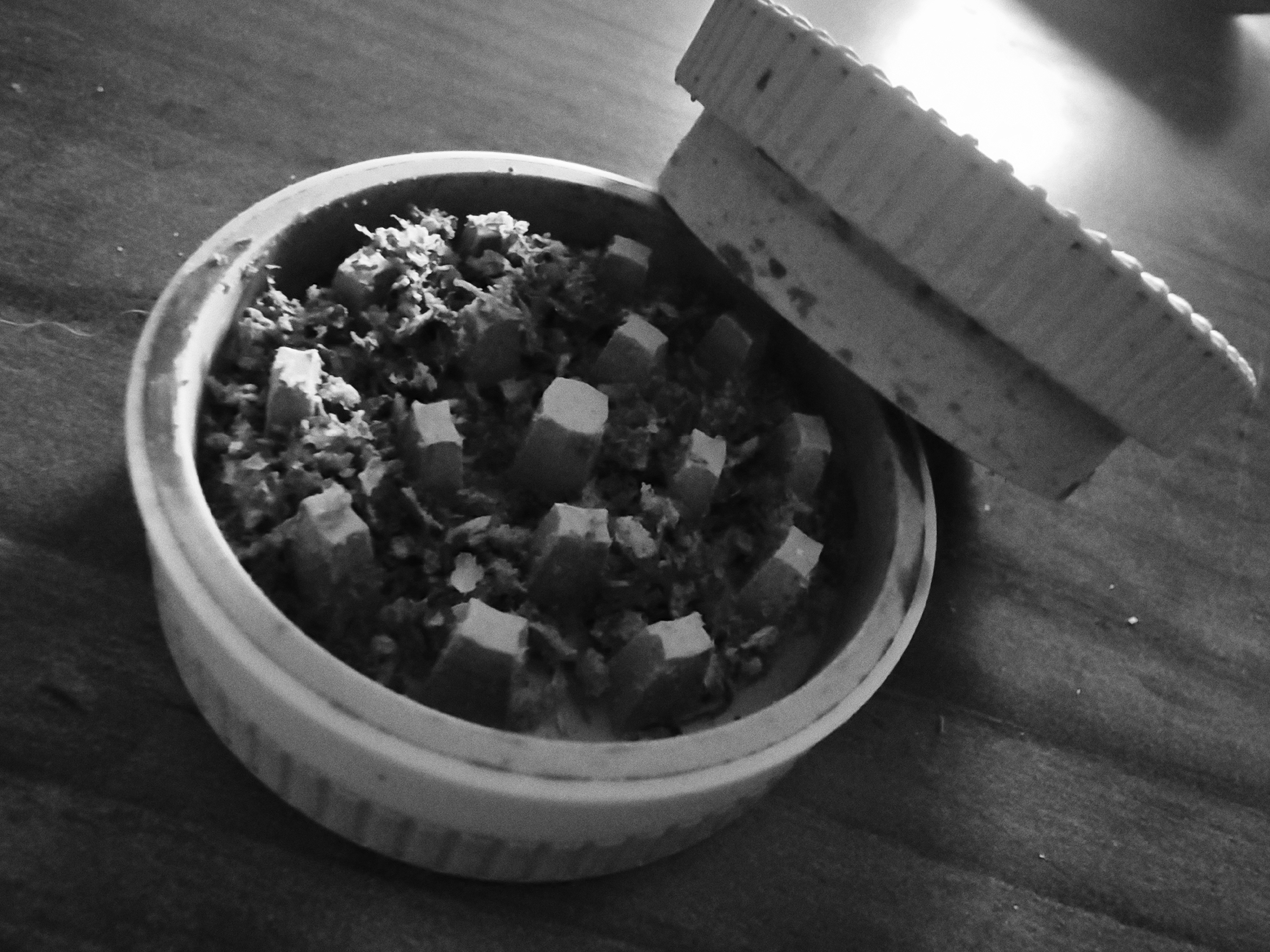 The image, in monochrome, shows a circular container with a lid partially covering its contents. The container appears to be made of a rigid material, possibly ceramic or plastic, with a ridged exterior design. Inside, there is a mixture of small, irregularly shaped pieces and cube-like objects, suggesting some form of processed or prepared substance. The lid resting to the side also exhibits a ridged design on its edge, matching the container. The background is a dark, textured surface, possibly a wooden table.
There is no discernible information in the image to suggest any location, including Varna, Bulgaria. The scene appears generic and could be anywhere. No people, specific actions, or interactions are visible.