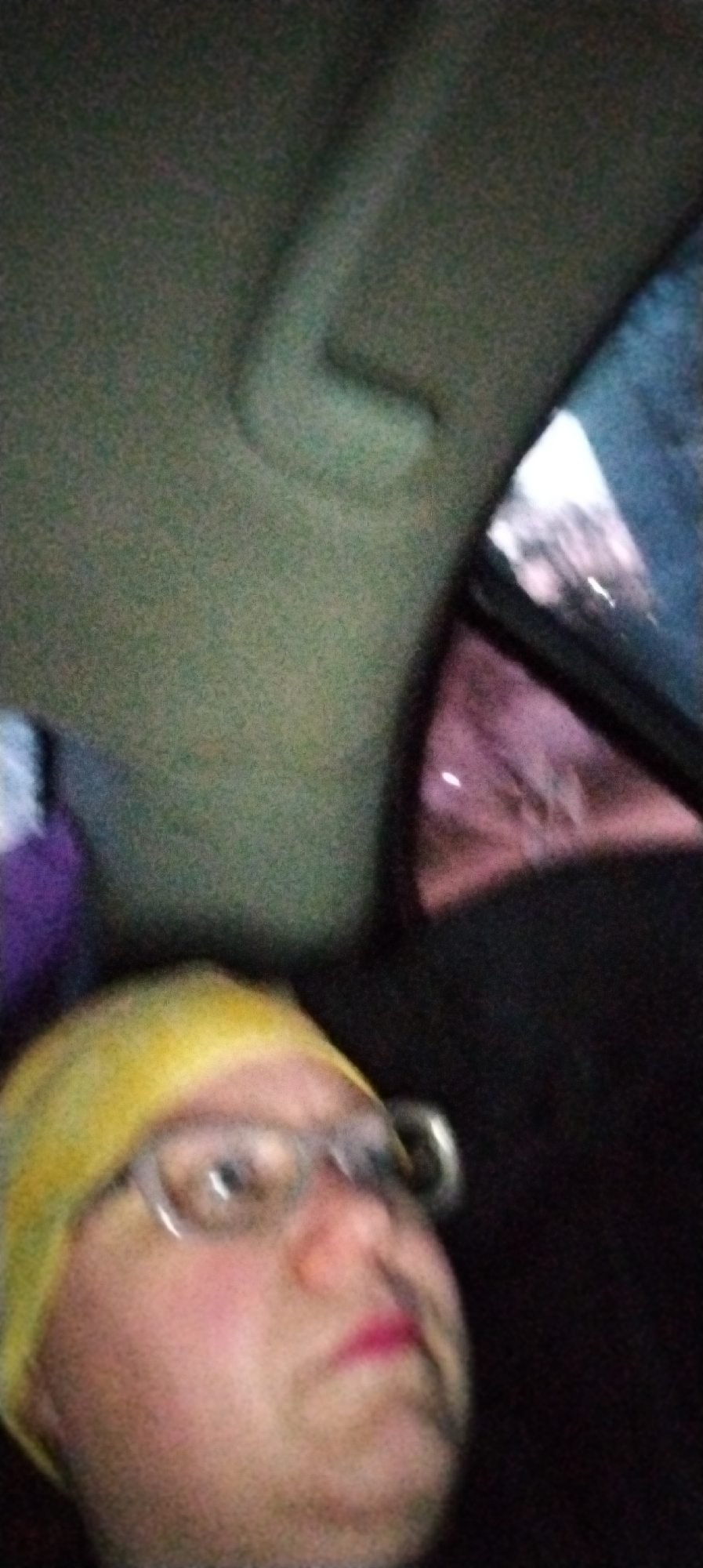 A blurred selfie depicts a person wearing a yellow beanie and glasses inside a vehicle, likely at night. A handle is visible in the upper left corner, suggesting it is a car's interior. Through the car window, city lights are faintly visible, suggesting an urban environment. The location is not identifiable from this image.