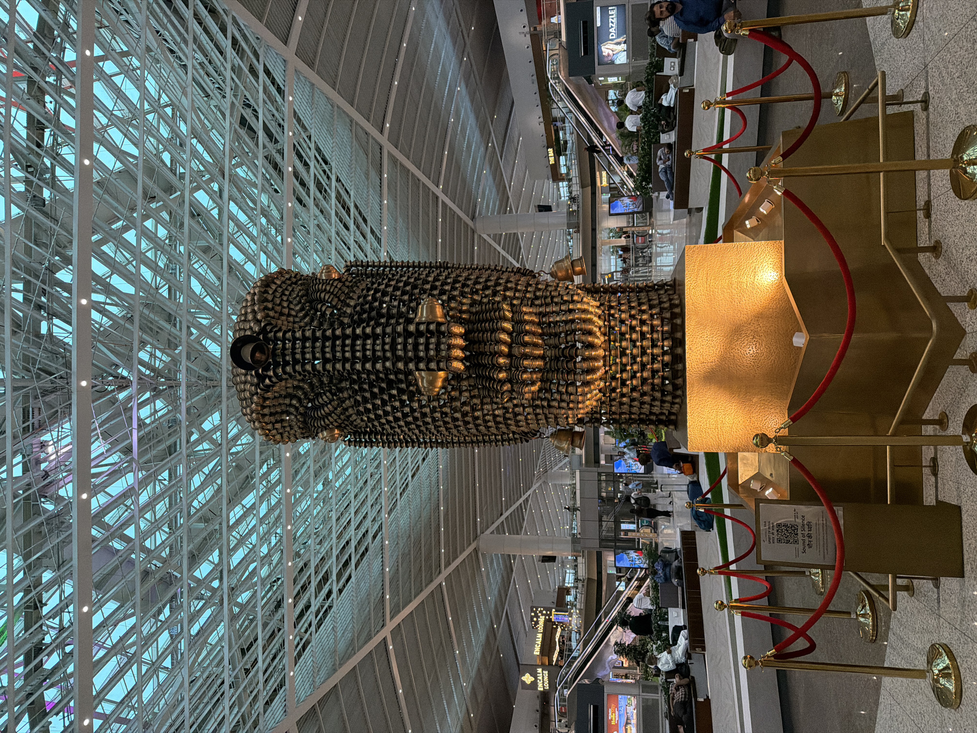 The image shows a large, ornate sculpture of a face, constructed from numerous small bells, dominating the center of the frame. The sculpture is illuminated from within, casting a warm glow. It is situated in a modern, spacious indoor environment that appears to be an airport terminal, characterized by a high, arched ceiling with a complex metal framework and translucent panels that allow light to filter through. Escalators and signage indicating "ENCALM LOUNGE" and "THE BAR" are visible in the background, suggesting a commercial and transit hub. Several people are present in the scene, some seated on benches or resting, while others are standing or walking. The sculpture is cordoned off with red velvet ropes and gold stanchions. A QR code and text in English and Hindi are visible on a sign near the sculpture, reading "Sound of Silence" and "मौन की ध्वनि". This setting is consistent with the Indira Gandhi International Airport (DEL) in New Delhi, India, known for its impressive art installations.