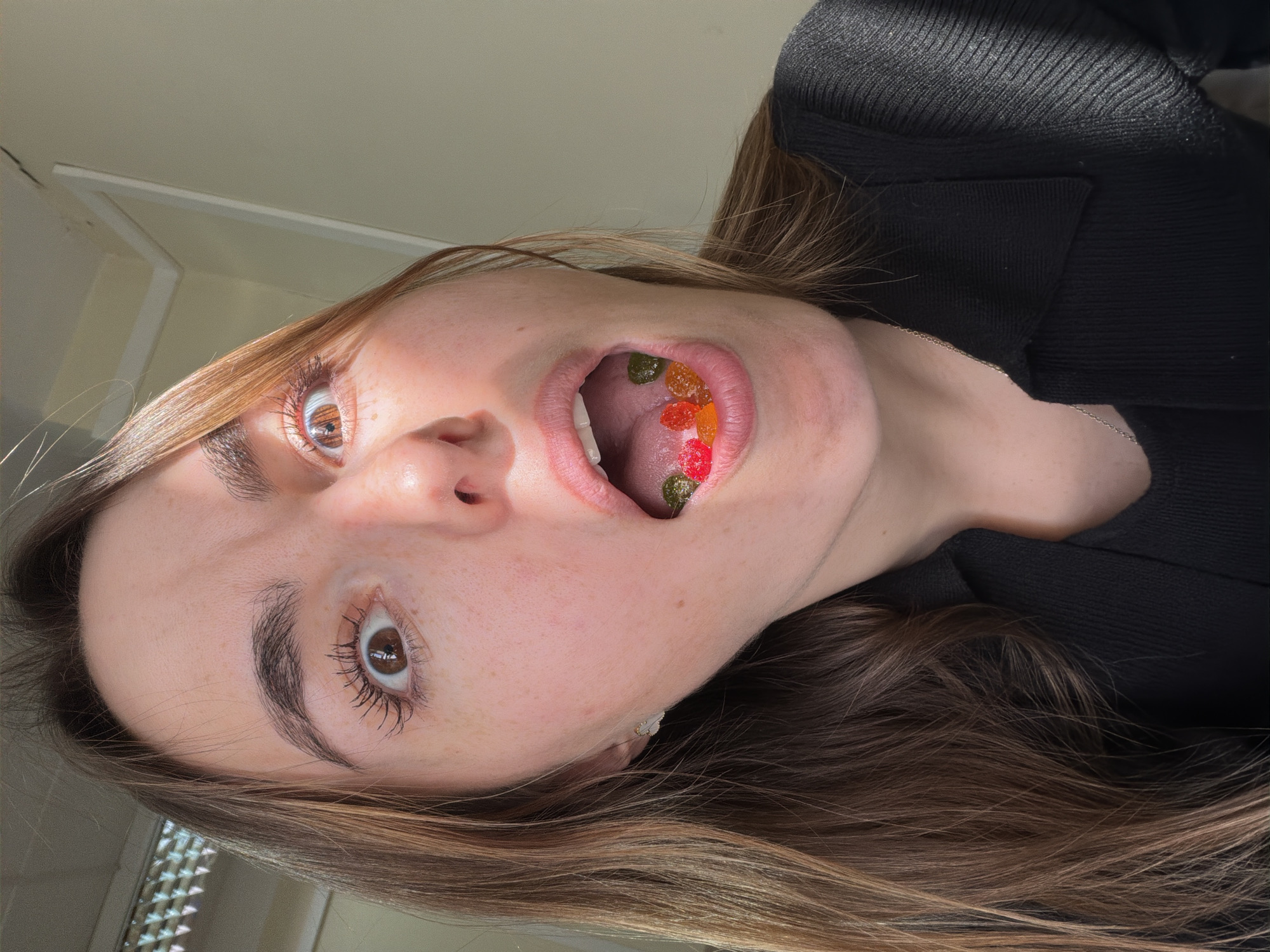 A young woman with fair skin, light brown hair, and brown eyes is holding her head back and looking directly at the camera with her mouth open, revealing several gummy candies inside. She is wearing a black, textured top. The background shows a light-colored wall with a hint of window blinds in the bottom left corner. The setting appears to be indoors. There is no indication that this image was taken in Izhevsk, Russia.