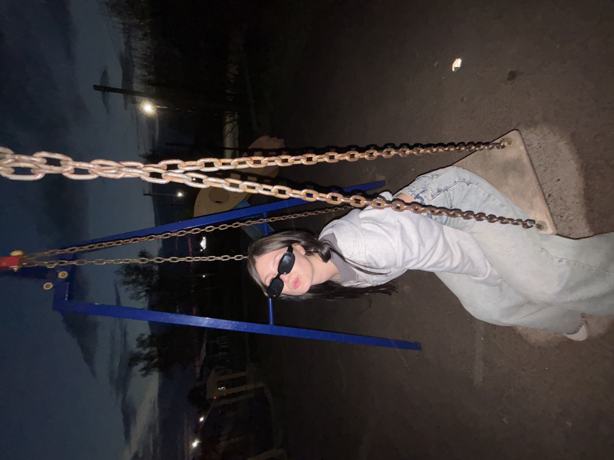 A young woman wearing sunglasses, a light-colored sweatshirt, and light-wash jeans sits on a swing set in a playground. The swing set is constructed of blue metal with chains. The background shows a dark, outdoor area with trees and faint city lights, consistent with an evening or night setting. The geographic location is given as Фоки, Russia.