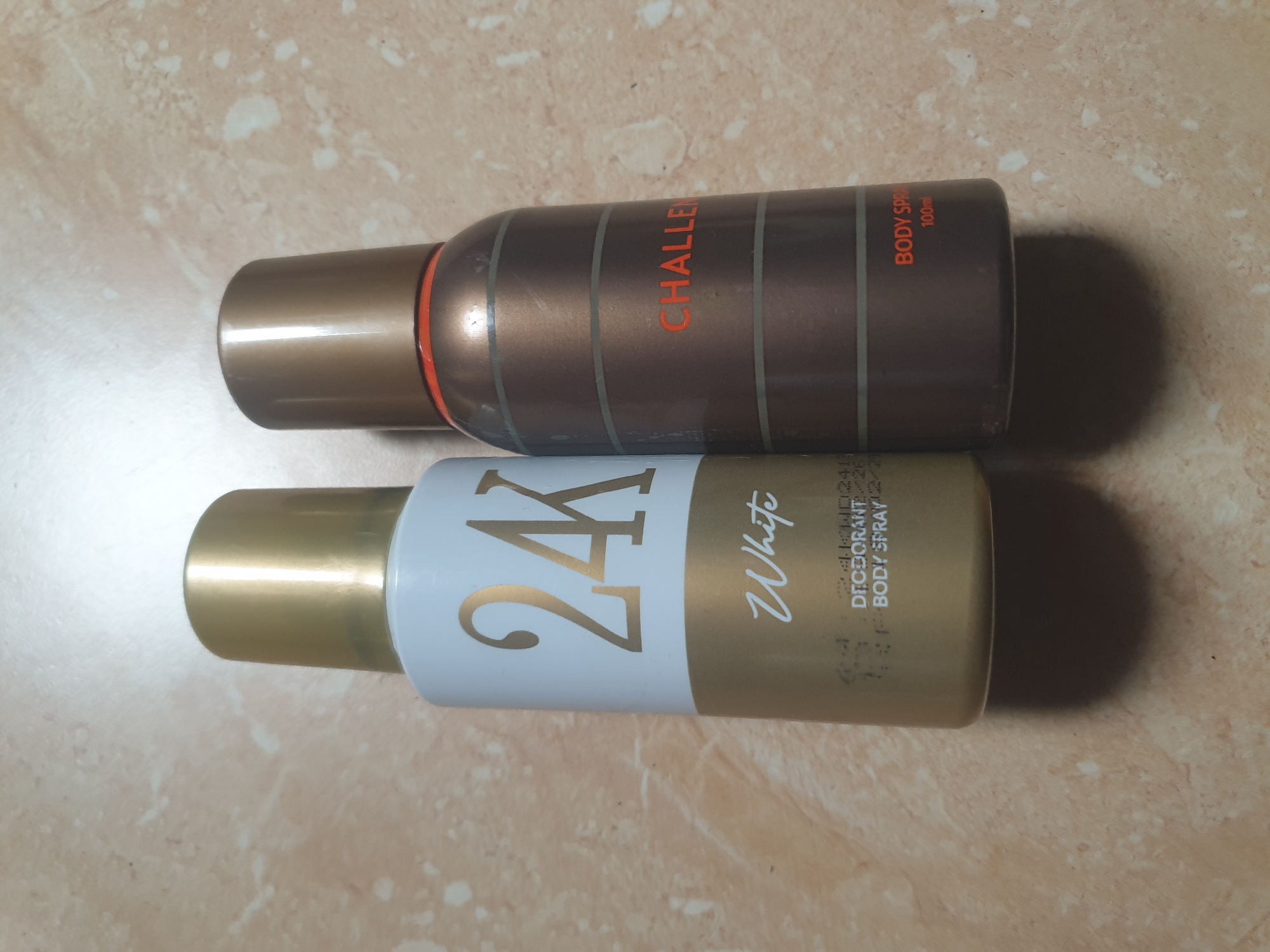 The scene displays two upright aerosol spray cans situated on a mottled, light brown and beige tiled or laminated surface, likely indoors in Jalingo, Nigeria. Overhead lighting casts subtle shadows behind both objects.
The left spray can features a metallic gold cap, a matte white upper body, and a metallic gold lower body. "24K" is emblazoned in large gold numerals and letters on the white section, with the word "White" in gold script directly below it. Towards the bottom of the gold section, faint black text is visible, including "DEODORANT" and "BODY SPRAY".
The right spray can has a metallic brownish-bronze cap and a matching body with a subtle sheen. A thin red band encircles the can directly beneath the cap. The word "CHALLENGE" is prominent in orange capital letters on the mid-section. Below this, "BODY SPRAY" and "100ml" are printed in smaller orange text. The can's body also exhibits faint, horizontal groove lines. No individuals are present in the image.