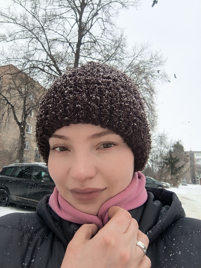A female individual is depicted in a close-up outdoor portrait against a winter backdrop in Vinnytsia, Ukraine. The subject, positioned centrally and facing forward, has light brown eyes and a neutral expression. She wears a dark brown, textured knit hat, lightly dusted with white snow particles, and a light pink neck garment beneath a dark, padded winter jacket. Her right hand, featuring a silver-toned ring on the ring finger, is positioned near her neck, appearing to grasp the jacket collar or scarf.
The background reveals a snow-covered urban environment under an overcast sky. Leafless deciduous trees are prominent, their branches stark against the pale sky. Multi-story residential buildings are visible; one on the left, an older structure with visible windows, and another further right. Parked automobiles, including a dark-colored station wagon or SUV, are present on a snow-lined street. Several dark-feathered birds are observed in flight in the upper right portion of the scene, while light snowfall is evident in the air and on surfaces.