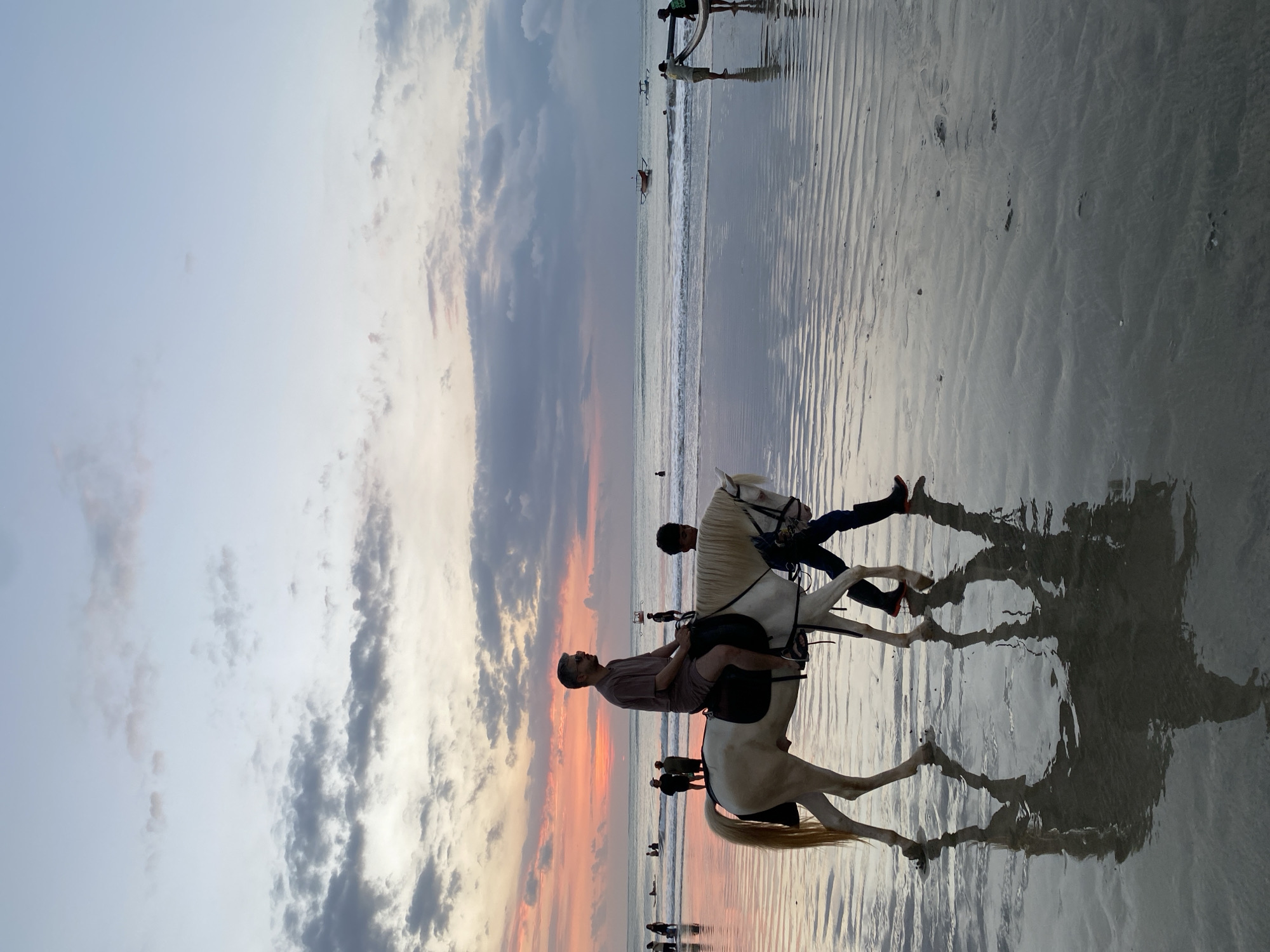 This image captures a vibrant beach scene at either sunrise or sunset in Kuta Selatan, Indonesia. The central focus is a man riding a white horse, alongside another white horse being led by a second man. The rider, wearing a dark shirt and sunglasses, is seated on the horse facing the ocean, while the handler, dressed in dark clothing, walks beside the second horse, holding its bridle.
The horses and people cast prominent, dark reflections on the wet, sandy beach, which shows distinct ripple patterns from the receding tide. In the background, the vast ocean extends under a sky ablaze with soft hues of orange, pink, grey, and light blue near the horizon, gradually fading to a lighter sky overhead. Scattered clouds are visible, adding texture to the sky.
Numerous other people are spread across the beach and in the shallow waters along the shoreline, some wading, swimming, or simply enjoying the seaside. Further out in the ocean, a few small boats or canoes can be faintly seen. The time of day is clearly denoted by the colorful sky and the low, golden light reflecting off the wet sand. The scene conveys a peaceful and recreational atmosphere typical of a tourist beach. No discernible text is visible in the image.