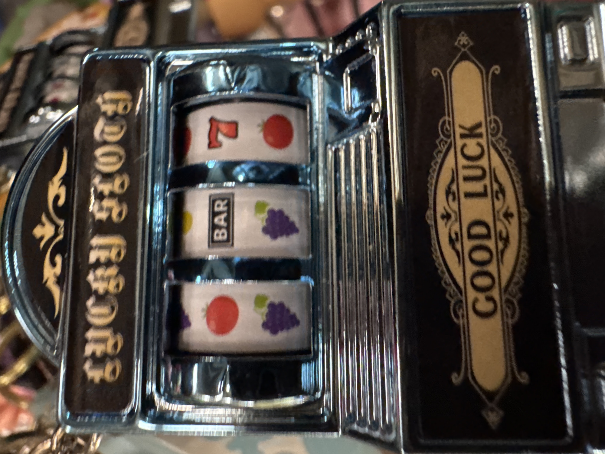 This is a close-up, high-angle shot of what appears to be a miniature slot machine or a slot machine-themed novelty item, possibly a lighter or a decorative box. The object is predominantly dark with shiny, chrome-like metallic accents.
The central part of the item features three visible reels, typical of a slot machine. The top reel displays a red '7' next to a red cherry or tomato. The middle reel shows the word "BAR" in white text on a black background, beside a cluster of purple grapes. The bottom reel also displays a red cherry or tomato next to purple grapes.
To the left of the reels, there is a decorative panel with golden, ornate text vertically oriented on a dark background, which reads "LUCKY STORY". To the right of the reels, another decorative panel features the words "GOOD LUCK" written vertically in gold letters within an elaborate, framed design on a dark background.
The metallic surfaces are reflective, showing hints of the surrounding environment, which appears to be indoors with artificial lighting. The object is stationary, and no activity is taking place. The focus is sharp on the slot machine details, with the background slightly blurred.